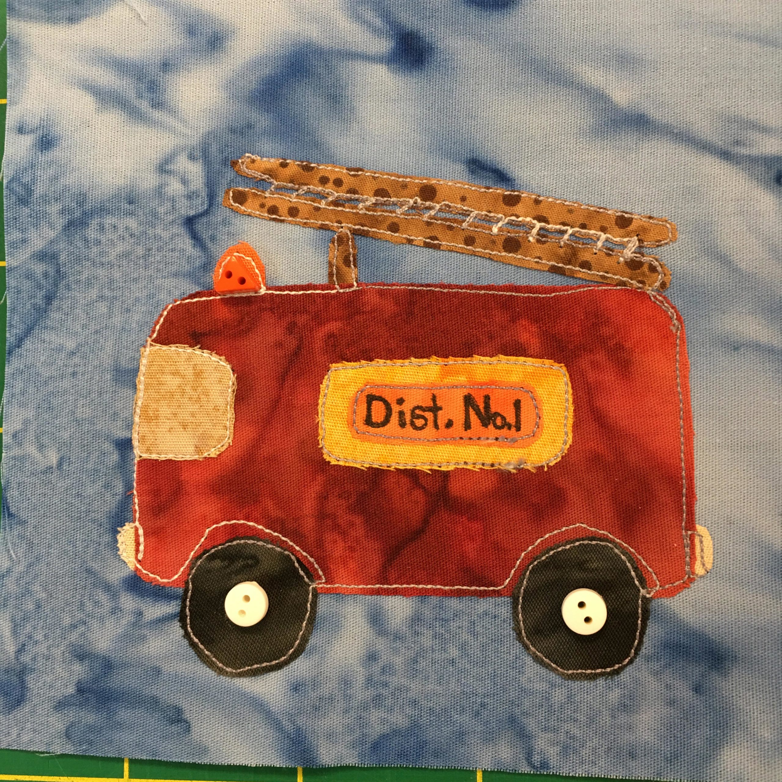 This quilt block shows a perfectly rectangular fire truck made of a deep red fabric. In the front the cab windshield is a splotchy golden yellow. As we look at it from the side we can see a orange sign bordered in yellow that says,