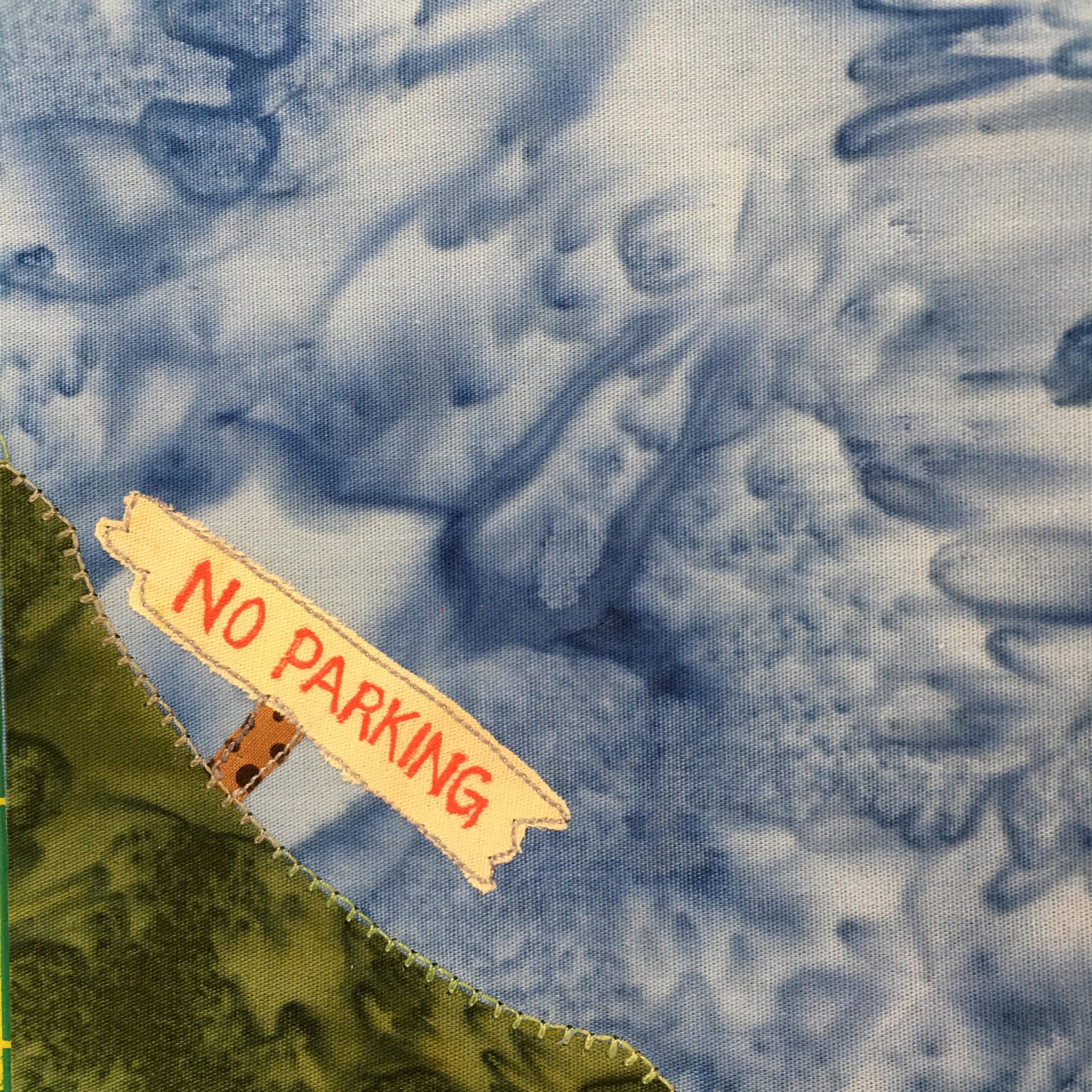 This block shows a corner block with a blue salmon-patterned background. There is a rich green grass patch with light yellow stitching in the bottom left corner. On the mount of grass fabric is a sign with an orange polka-dotted stick holding a light yellow sign with red lettering on it that reads, 