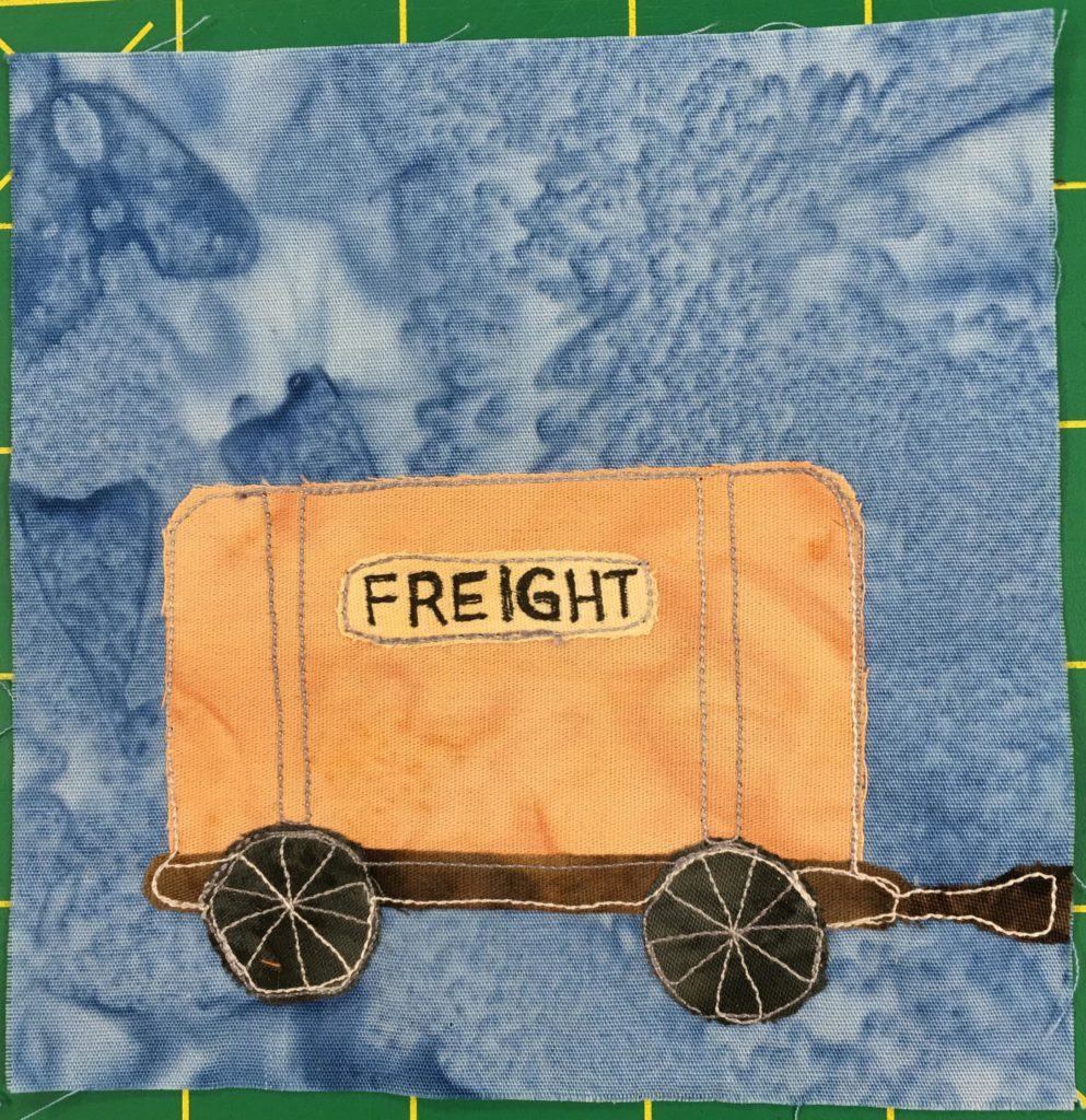 This quilt block shows a railroad car against a light blue salmon-patterned background. The railroad car is a light orange box with a yellow sign on the side with black lettering that reads,