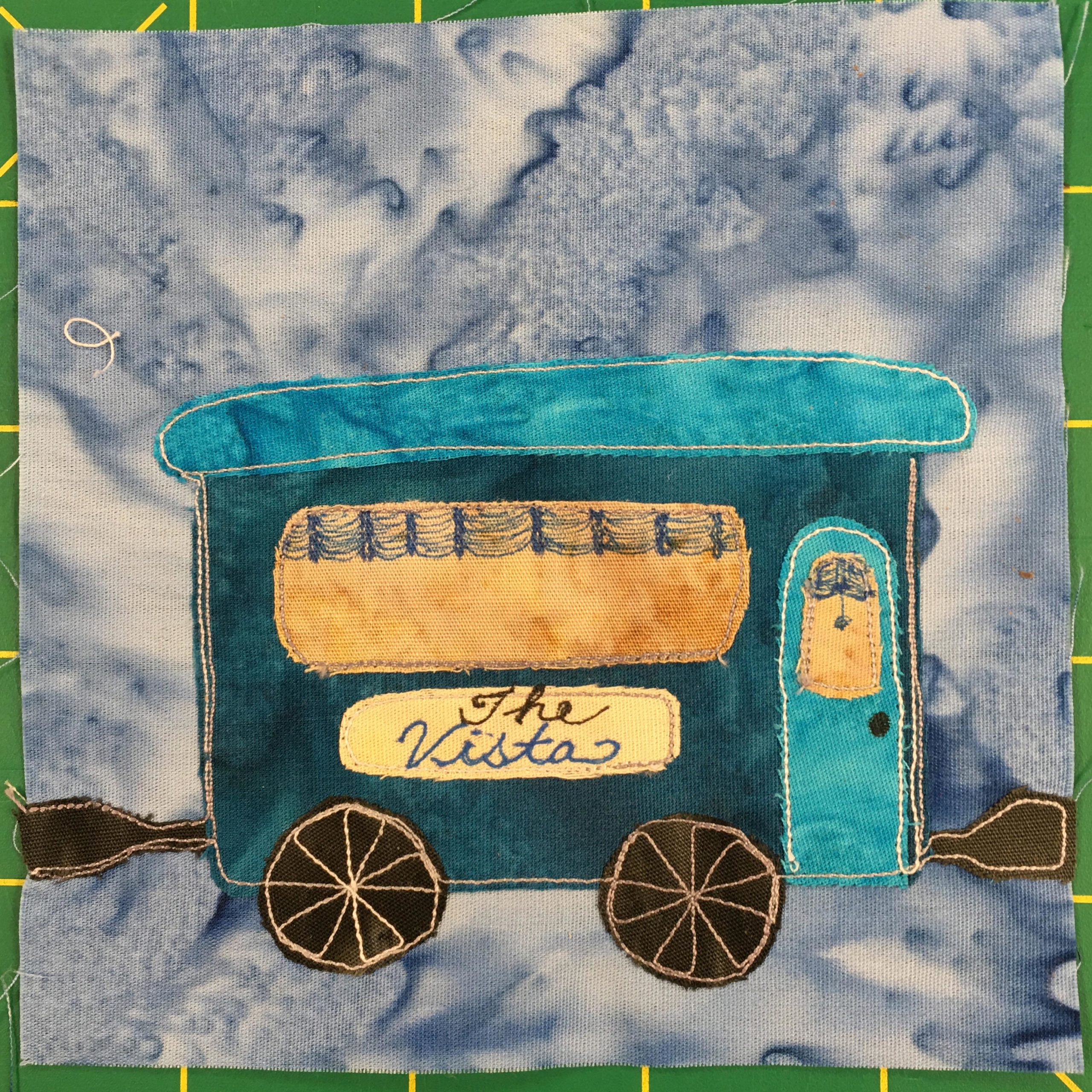 This quilt block shows a very blue railroad car against a faded blue salmon-patterned background. The roof is a bright light blue as well as the door. The rest of the rectangular car is a darker blue. The windows, one in the door and a large single window on the side are a dark orange-yellow. There are drapes in the windows that are pulled up defined by a blue pen drawing. There is a sign underneath the larger window that reads in cursive, 