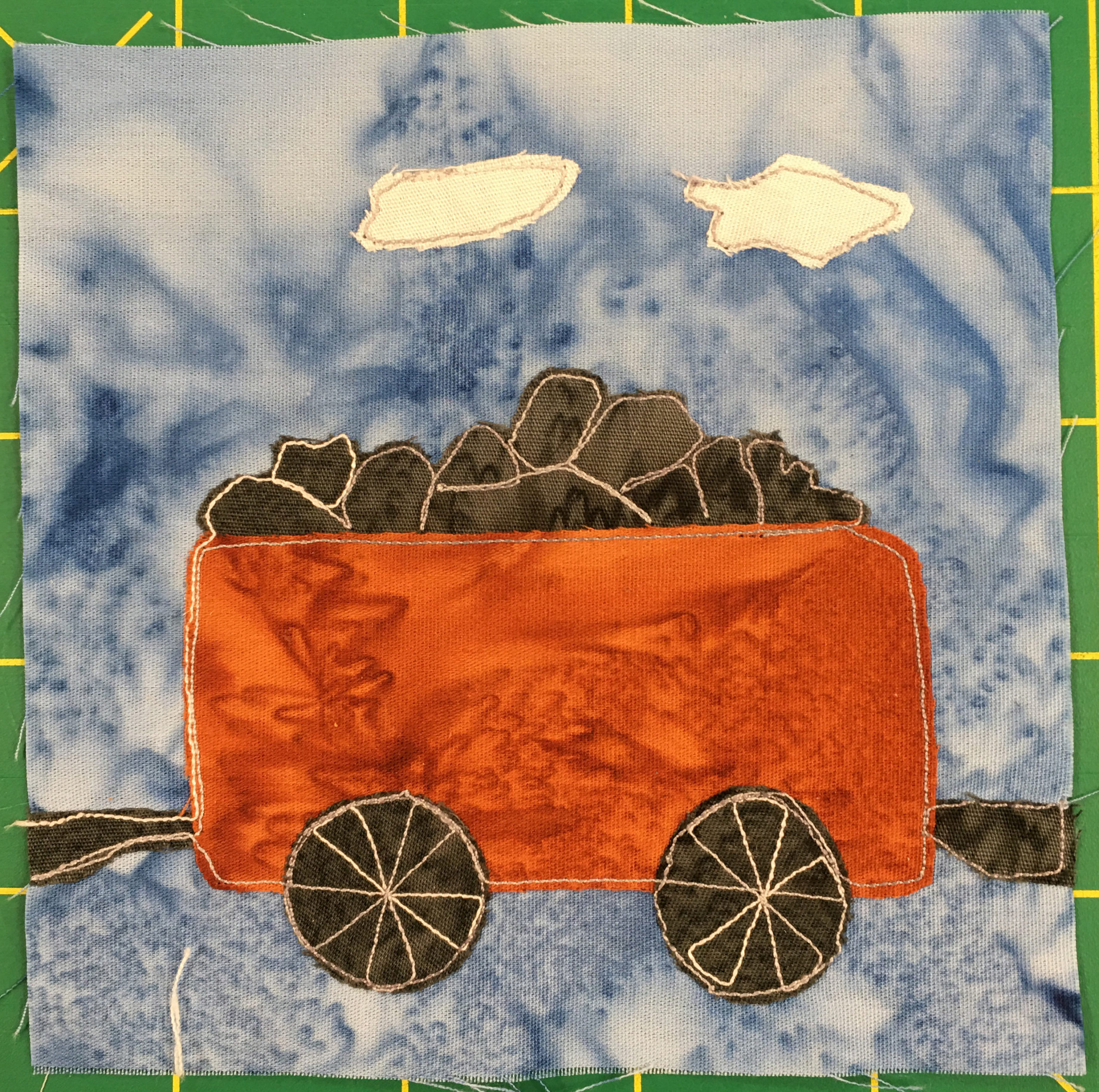 This quilt block shows a coal railroad car against a light blue salmon-patterned background. There are two white puffs above the coal car indicated with white fabric (from the engine). The coal is a dark fabric with light yellow stitching indicating the coal piles. The car itself is a deep orange. Two dark-brown wheels, the same color as the coal, are below the car with the spokes outlined in a similar light yellow stitching.