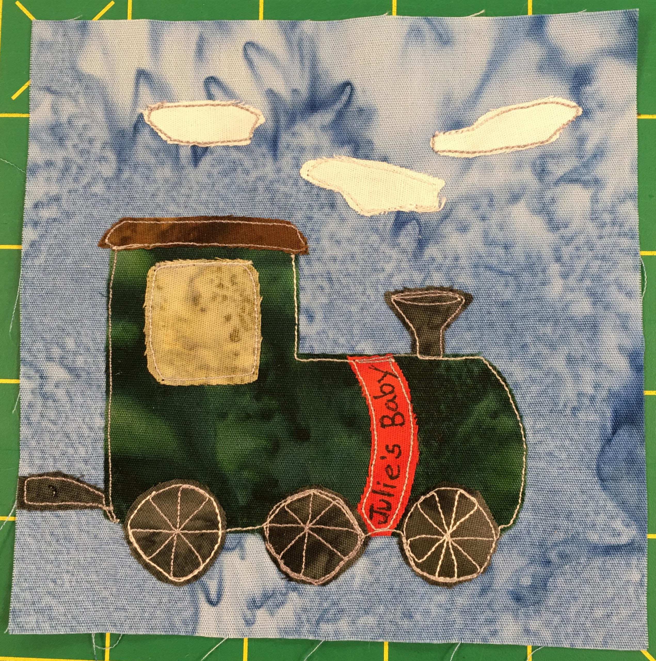 This quilt block shows a dark green train engine from the side against a light blue salmon-patterned background. There are three white puffs of smoke represented by white fabric above the smoke stack in the front and wafting behind. The roof of the cab is a dark brown while the window of the cab is dark yellow. There are three dark brown wheels with stitched spokes below. But most important is a ribbon of red around the engine that reads