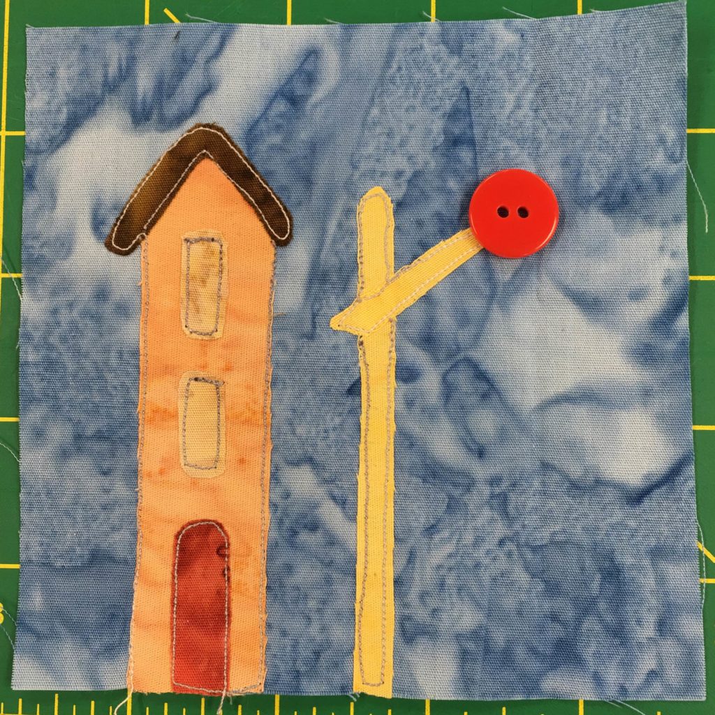 This quilt block shows a railroad crossing signal and building against a light blue salmon-patterned background. The building next to the signal is tall and orange with a small red door. The signal itself is bright yellow with a large red two-holed button at the end of it indicating the light at the signal.