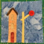 This quilt block shows a railroad crossing signal and building against a light blue salmon-patterned background. The building next to the signal is tall and orange with a small red door. The signal itself is bright yellow with a large red two-holed button at the end of it indicating the light at the signal.