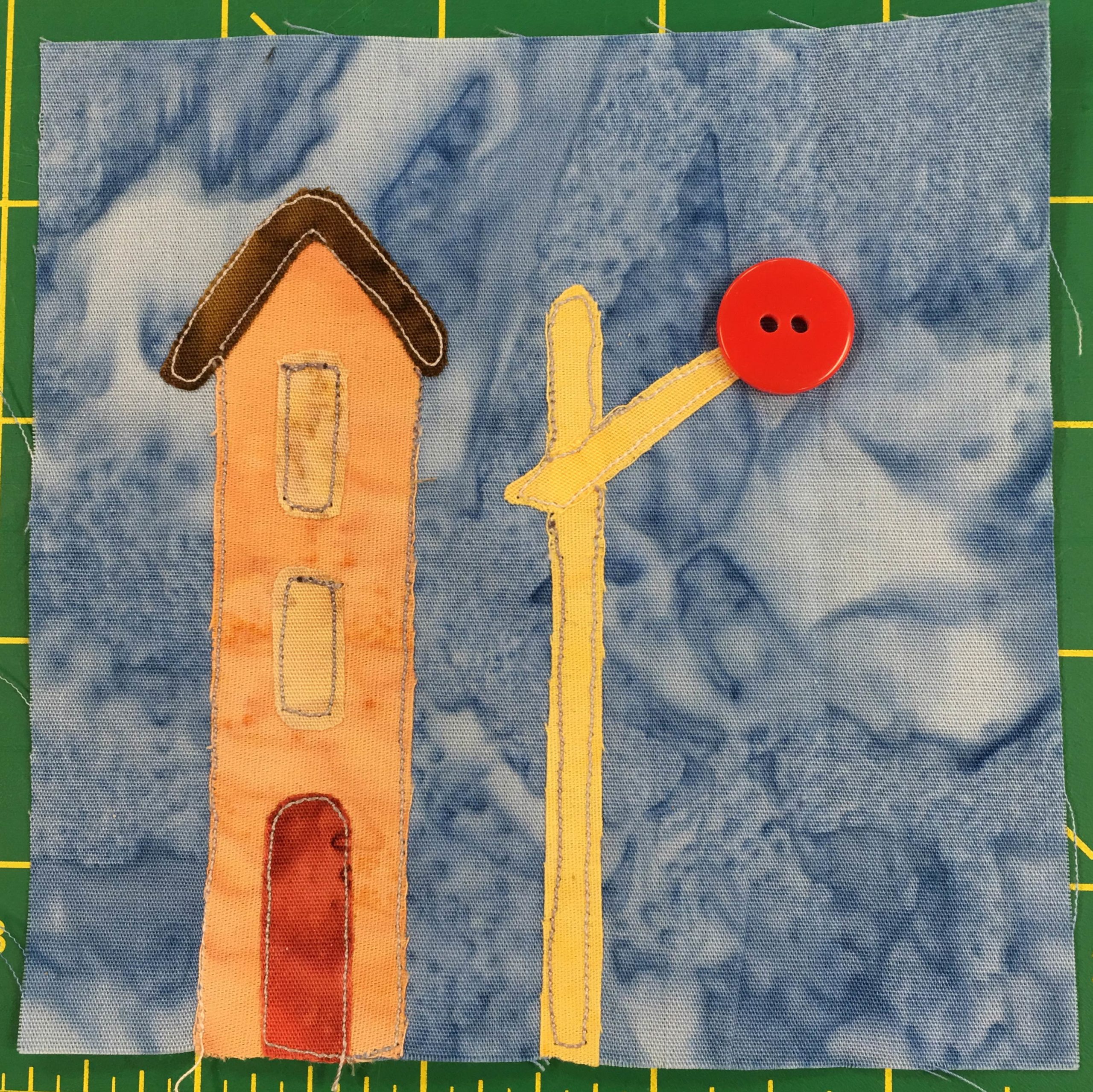 This quilt block shows a railroad crossing signal and building against a light blue salmon-patterned background. The building next to the signal is tall and orange with a small red door. The signal itself is bright yellow with a large red two-holed button at the end of it indicating the light at the signal.
