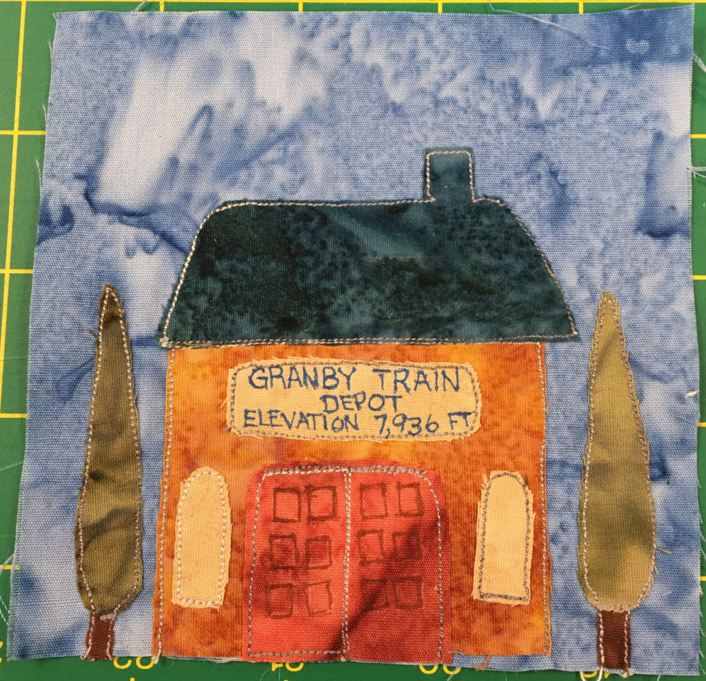 This quilt block shows a train depot with two poplar trees on either side. The train depot's roof is a dark blue with a little smoke stack coming out on the right. The roof itself is a large trapezoid shape. The building below is a bright orange square. Two large red doors are in the center with squares on them, and to either side of those doors are light yellow windows. Above the large double doors is a sign that reads in blue against light yellow, 
