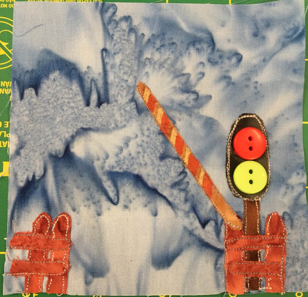 This quilt block shows a railroad crossing against a light blue salmon-patterned background. There is the end of an orange fence on the left, and in front of the crossing signal on the right is another piece of orange fence. The crossing signal has a brown post and at the top are two large two holed buttons, one bright red and one bright yellow, indicating the lights. There is a striped diagonal lighter brown, yellow, and orange strip of fabric coming out of the post at an angle representing the guard that lowers to stop cars from going on the tracks.
