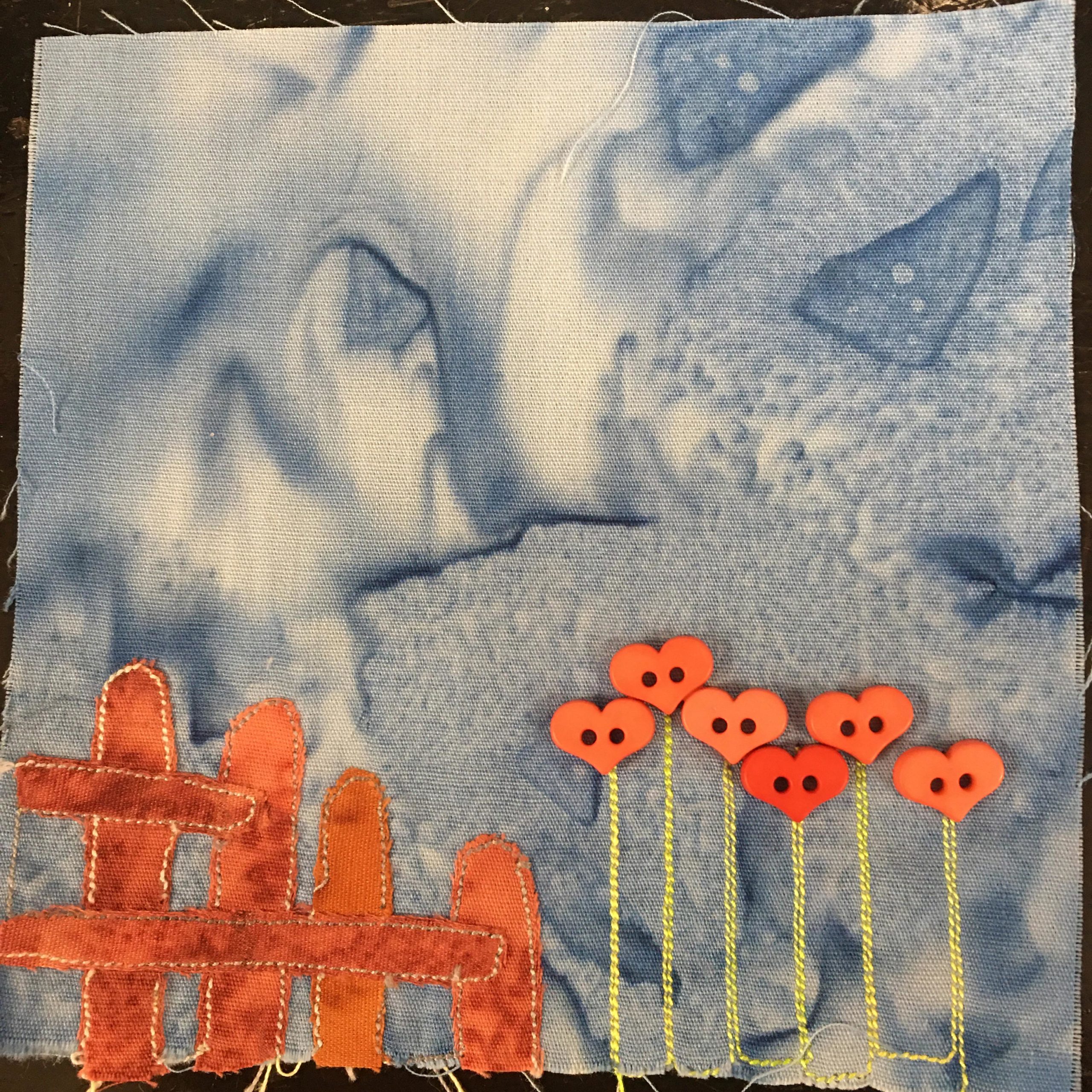 This quilt block shows the end of a bright orange fence with some flowers next to it against a light blue salmon-patterned background. The flower stems are yellow stitching and the flower bulbs are heart-shaped light red two-holed buttons.