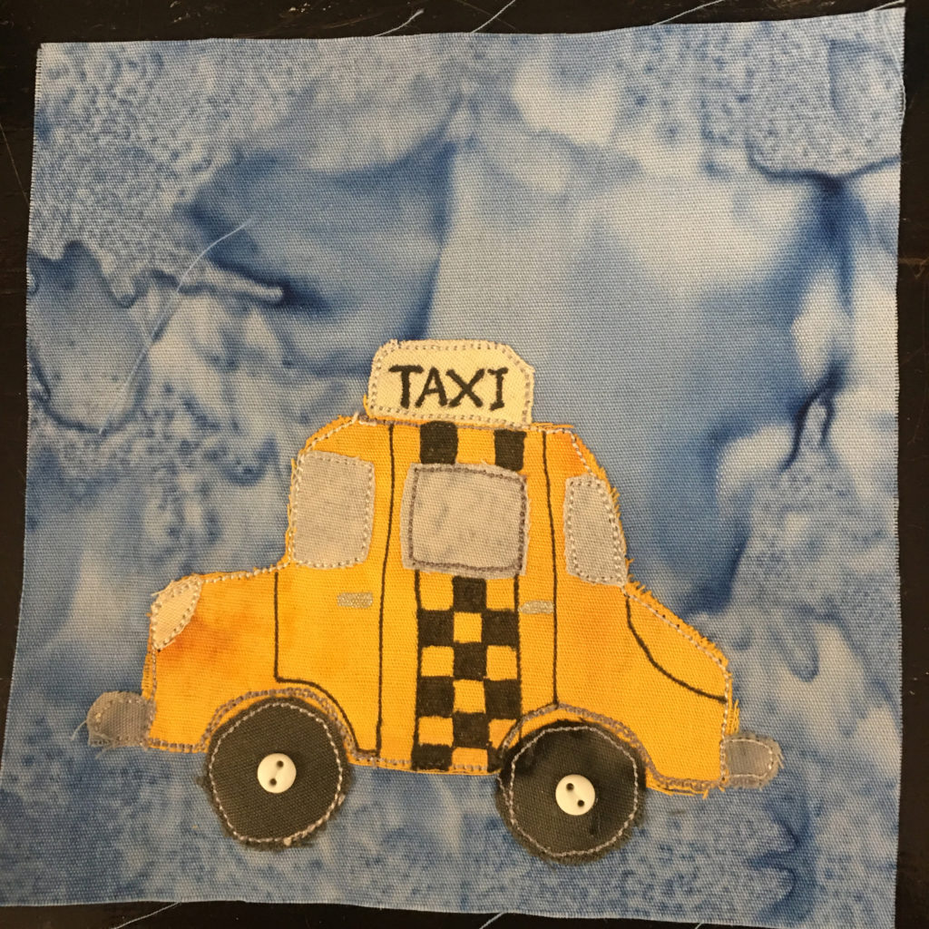 This quilt block shows a yellow taxi cab from the side against a light blue salmon-patterned background. The taxi is a bright yellow fabric, with light gray fabrics for the windows, windshield, and fenders. There is a light yellow sign on top that has the word