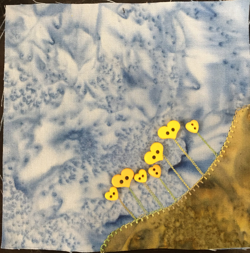 This quilt block shows a patch of grass in the bottom right that's a bit of a yellowy green tint of fabric. On the patch of corner fabric are straight stitched lines representing stalks of flowers. The flower buds are little yellow heart shaped two-holed buttons. This is all against a light blue salmon-patterned backgorund.