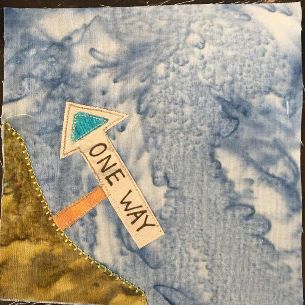 This quilt block shows a patch of grass in the lower left corner of the quilt block. This patch is a somewhat yellow green, mostly green, fabric color. On the patch of grass, tilted and going towards the center of the square is an arrow sign pointing left on a post. The post is orange, and the sign is white with a blue triangle at the end inside the arrow. On the sign are written the words,
