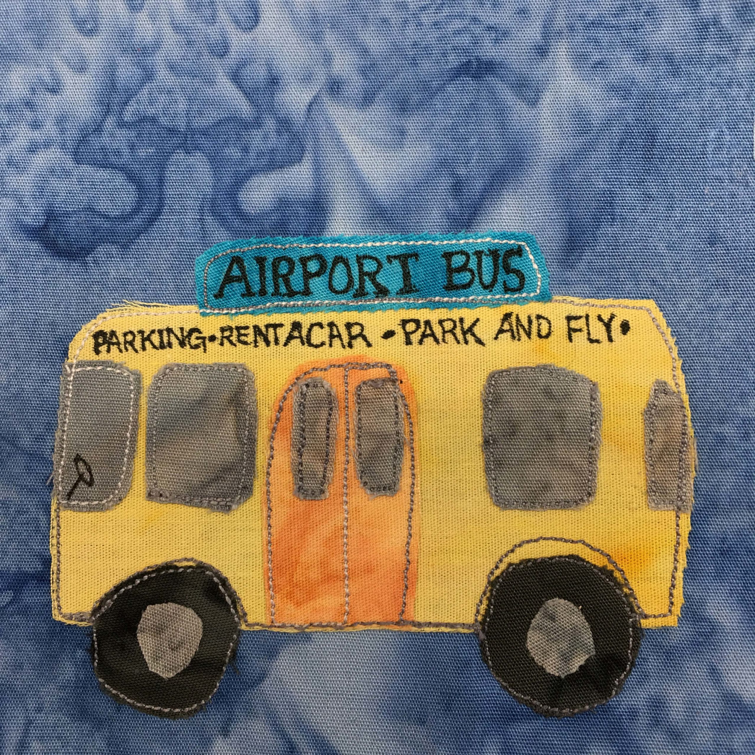 This quilt block shows the side view of an airport shuttle bus against a blue salmon-patterned background. The shuttle bus is yellow with gray colored windshield and windows. In the front windshield you can see in pen a steering wheel. On the side in the middle is an orange sliding double door. Above the windows and door in pen are the words, "Parking • Rent-A-Car • Park And Fly" and above that a blue sign oi top of the bus that reads in big capital letters, "AIRPORT BUS" There are two big black wheels at the bottom with gray patches of fabric inside indicating the hubcaps.