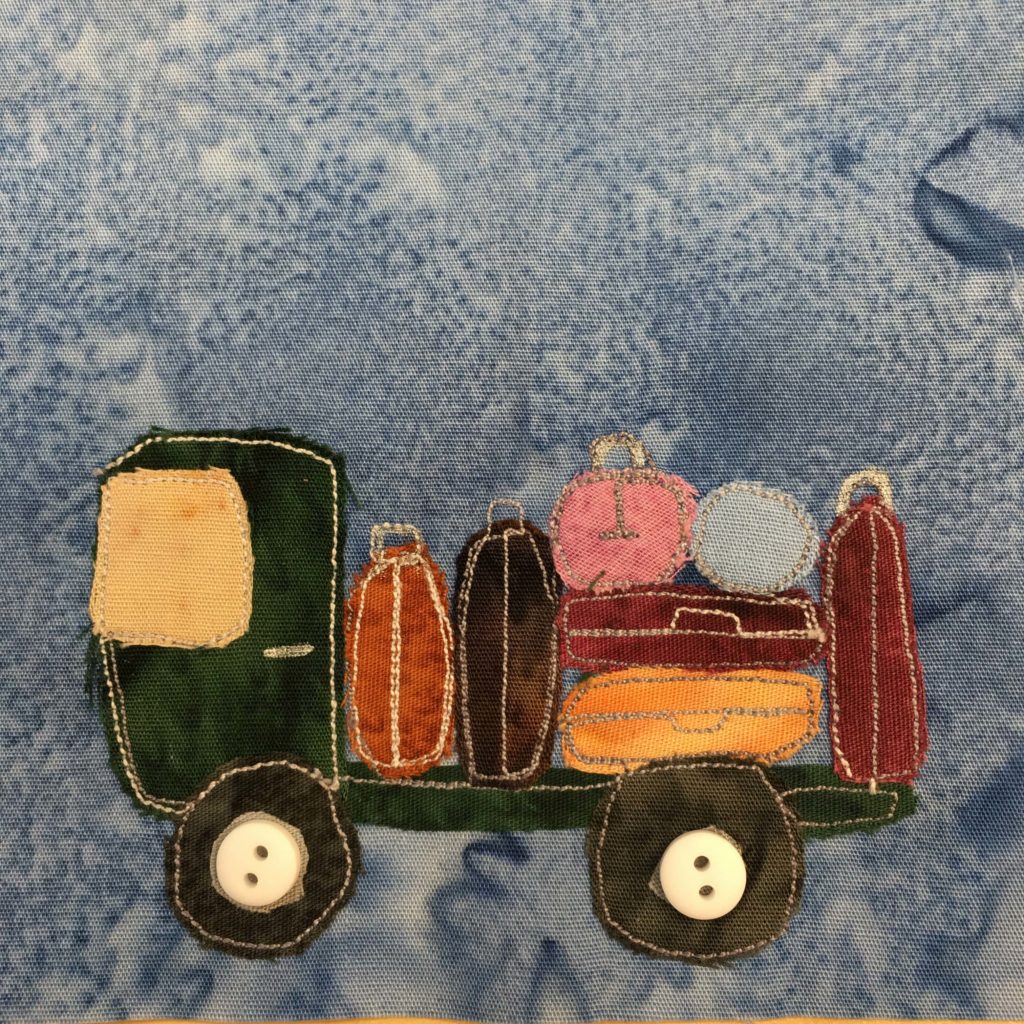 This quilt block shows a green luggage truck from the side against a blue salmon-patterned background. The truck itself is just a flatbed, and is made up of a dark green fabric. The cab has a yellow fabric windshield, and on the flatbed behind it are various suitcases. Each suitcase is a different shade of orange and brown, with one pink and one blue suitcase. There are two dark wheels below with fairly large white two-holed buttons on them indicating the axles or hubcaps.
