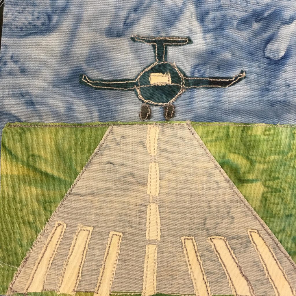 This quilt block shows a plane landing on a runway from the front. It's quite dramatic. The plane itself is a dark emerald blue with white stitching outlining it. The windshield is a patch of white fabric. The runway is a strip of gray fabric on green with white stripes of fabric showing the lines. Above the runway is a blue salmon-patterned background.