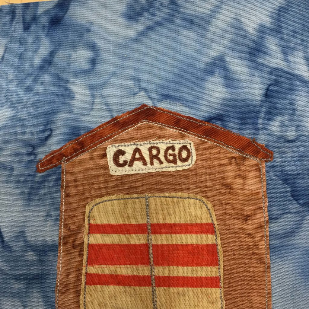 This quilt block show a very large building, much like an airport hangar. We're looking at the building from the front. It's very tall with a brown front, and a darker brown angled roof. Beneath the sharp apex of the roof is a light sign that has written on it in bold, "CARGO." Beneath the cargo signage are two large double garage doors (or one big door with stitching down the middle) that has three big red stripes across it horizontally. All of this is set against a blue slamon-patterned background.