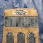 This quilt block shows a tall building with arched entryways on the bottom and a large span of vertical windows across the upper part. Above the windows is a sign that reads in blue pen, "Homer Municipal Airport." The windows are a gray fabric, with markings to show they reflect, and stitching showing the pane's edges. The archways below also have stitching. The building itself is a light tan fabric in the same salmon-pattern as the background which is blue.