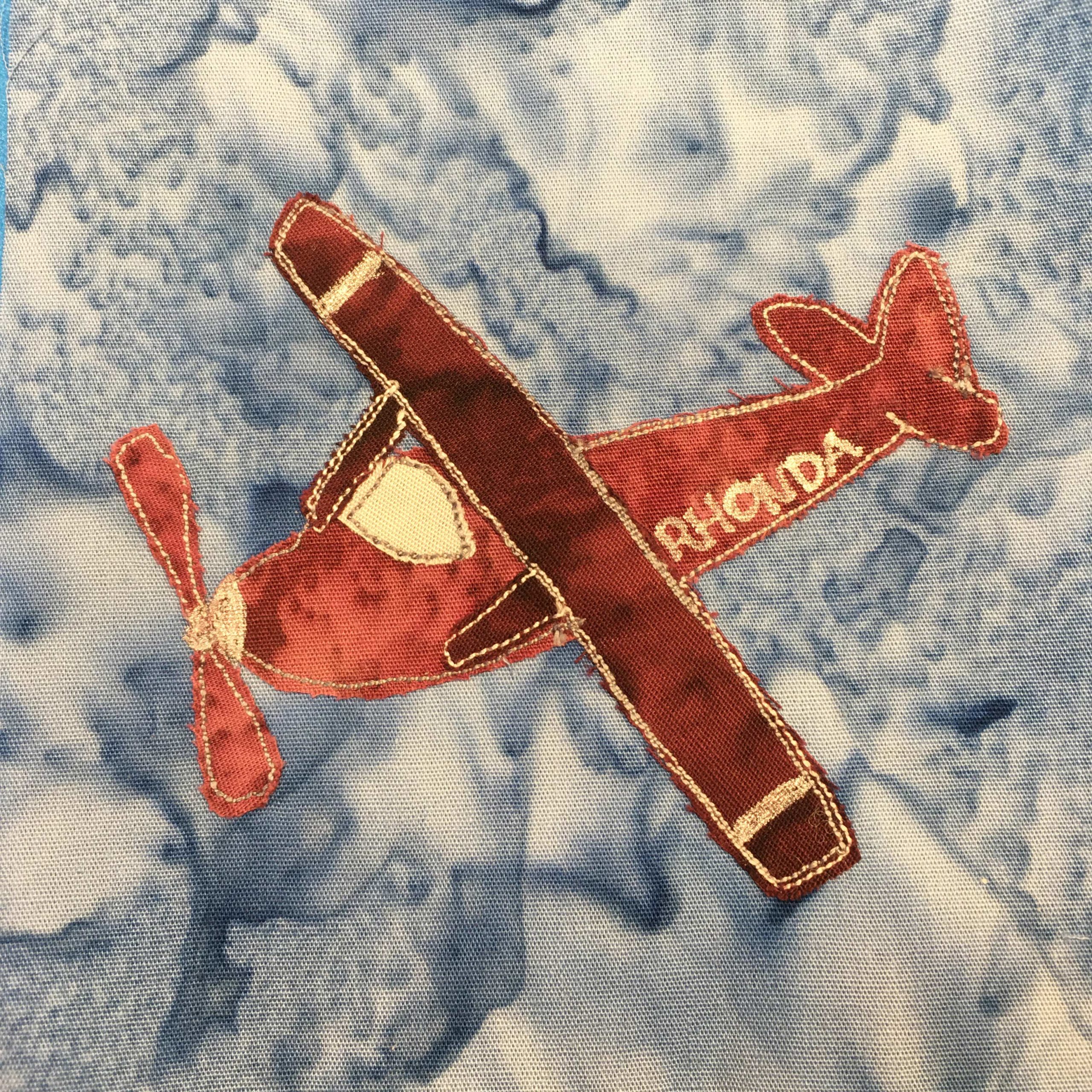 This quilt block depicts a deep red airplane as seen in the sky from an angle above. The whole airplane is a deep maroon red, with yellow stitching around the outlines. There is a windshield which is the same yellow color with red stitching on its outline. The plane has one wing above the hull, and one propeller blade on the end of its nose. On the side of the plane in yellow is the word, "Rhonda." This is all against a blue salmon-patterned background.
