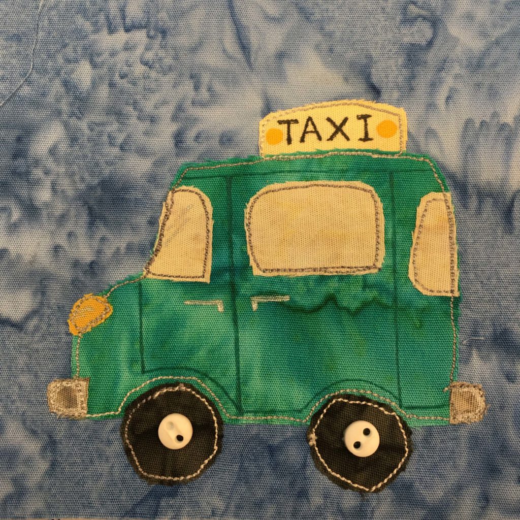 This quilt block shows a green taxi cab with a yellow sign on top that says "TAXI" in black pen. There are two yellow dots next to each side of the word. The taxi itself is more like a little buggy or jeep, and is entirely a deep green. The headlight is a bright yellow. It looks a bit like an old car from history. The wheels are black and round with white stitching on the outer edge and in the middle two ivory colored two-holed buttons indicating the hubcaps.