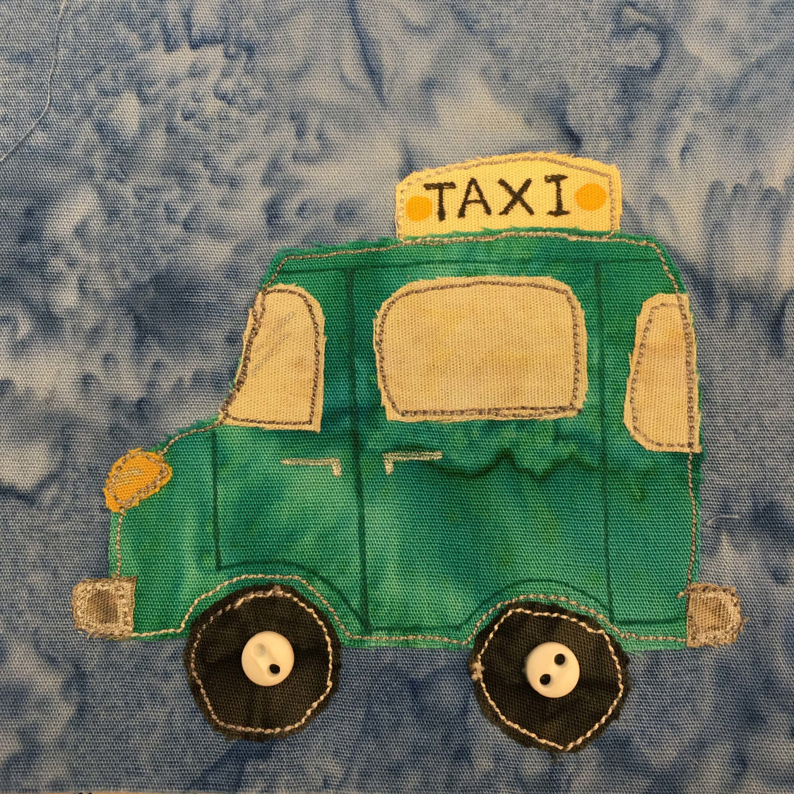 This quilt block shows a green taxi cab with a yellow sign on top that says "TAXI" in black pen. There are two yellow dots next to each side of the word. The taxi itself is more like a little buggy or jeep, and is entirely a deep green. The headlight is a bright yellow. It looks a bit like an old car from history. The wheels are black and round with white stitching on the outer edge and in the middle two ivory colored two-holed buttons indicating the hubcaps.