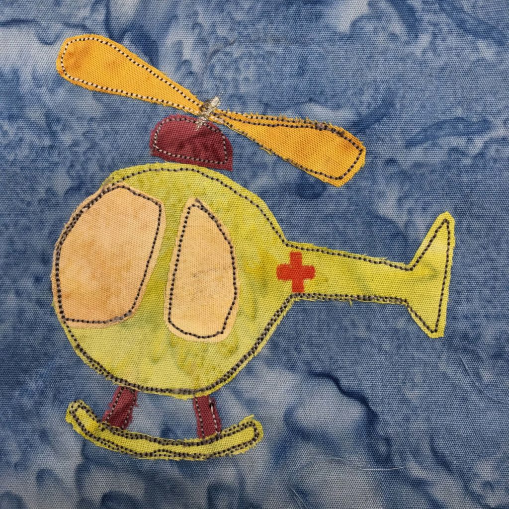 This block shows a round globular green helicopter with a first aid red cross on it. The propellers are yellow, and there are two large blades above the bubble. Below that is a deep red beanie cap like attachment that attaches the blades to the globe like body. The front of the ball has a large light yellow windshield, with a side door window next to it. This is all against a blue salmon-patterned background.