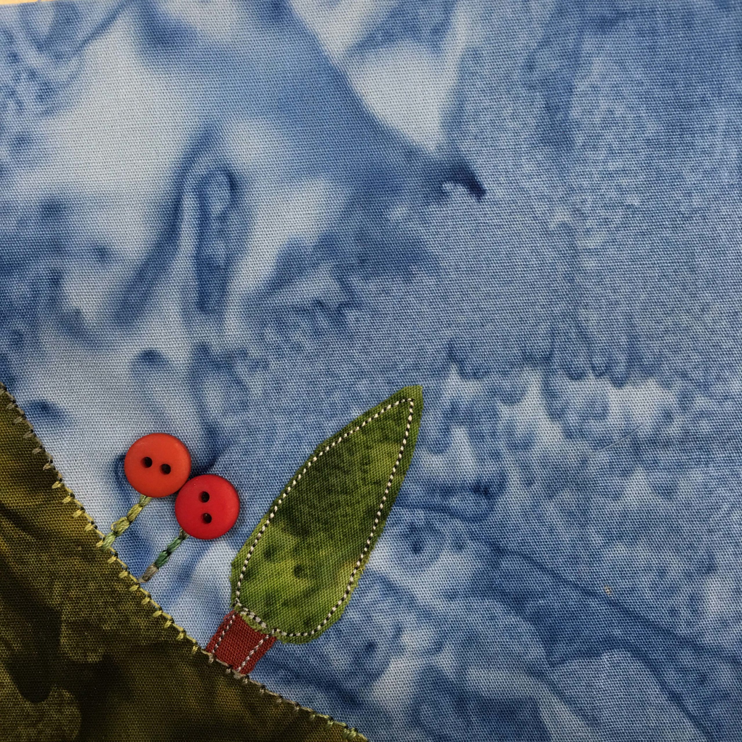 This quilt block shows a very dark patch of green fabric in the bottom left corner indicating a patch of green grass. On top of the curved patch of grass is a small tree, and two large red flowers (shown using small red two-holed buttons). There is lighter green stitching indicating the flower stalks and on the edge of the grass. There is also white and black stitching around the tree. Against this is a darker blue salmon-patterned background.