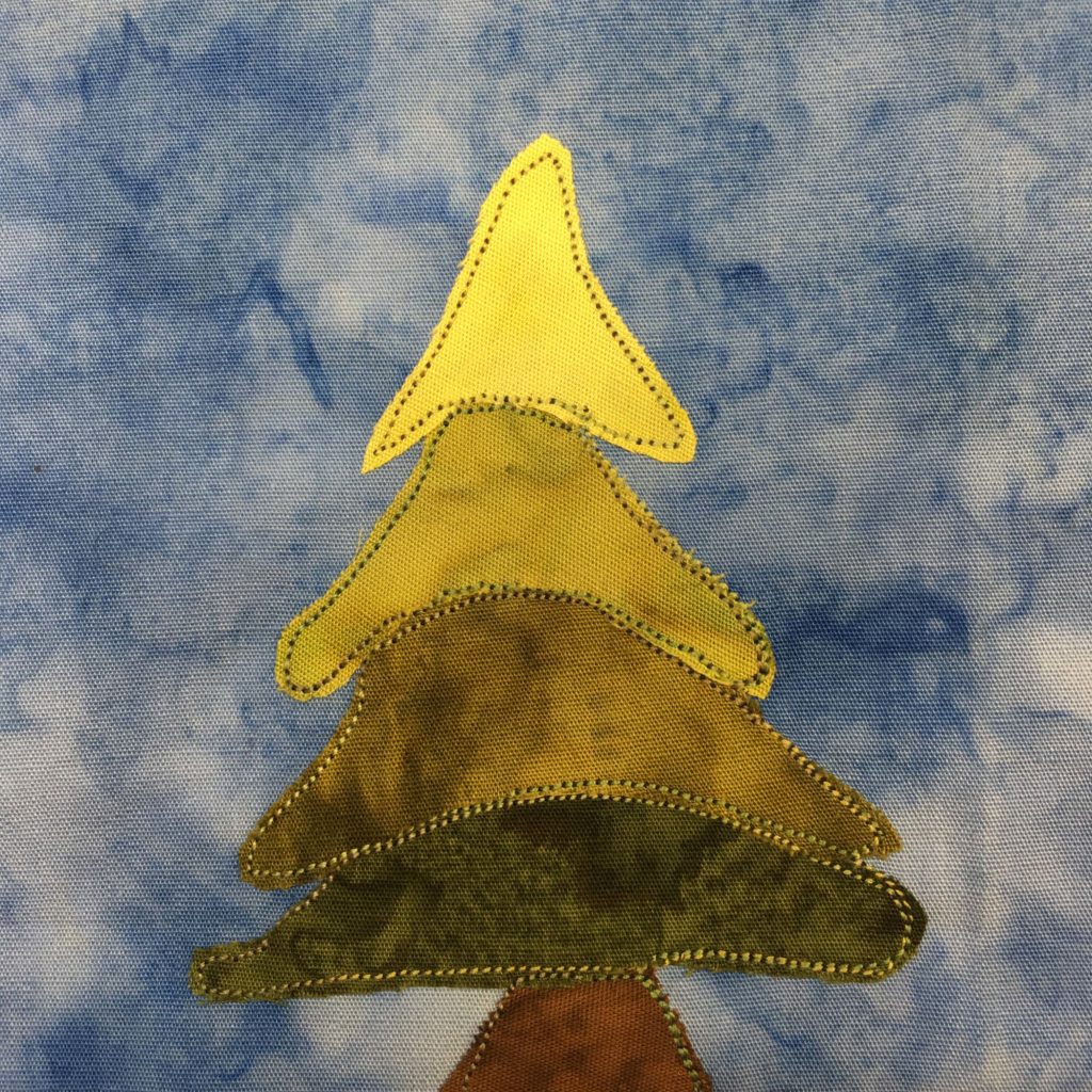 This quilt block shows a big evergreen tree. Each section from bottom to top is a lighter shade of green, starting with a dark green fabric. Each section, shaped a bit like a floppy triangle, is outlined by a dark green thread, including the trunk at the bottom which is a brown fabric. This is all against a blue salmon-patterned background.