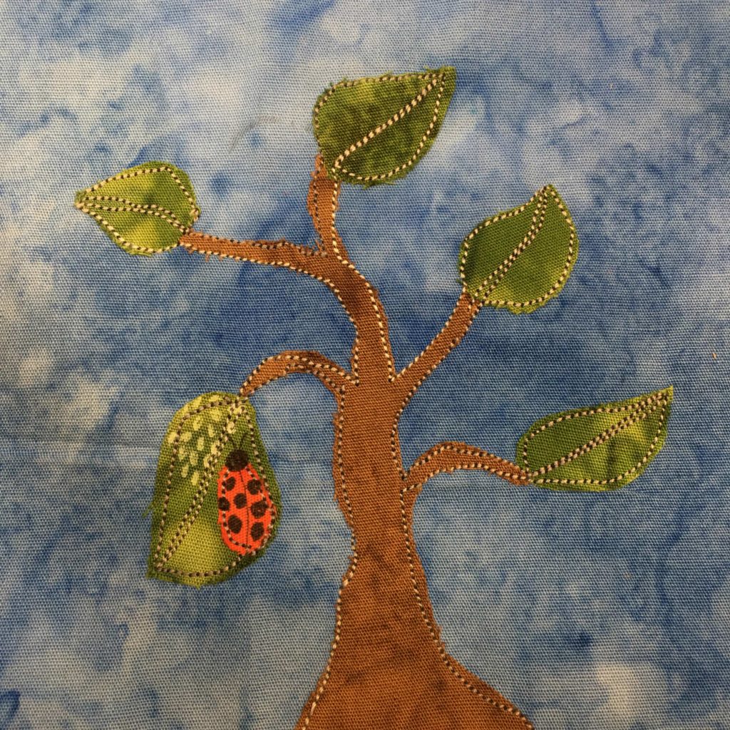 This quilt block shows a plant with a brown trunk and green leaves. The branches and singular leaves jut out in various directions. On one leaf on the left hanging down is a large ladybug with seven dark spots on its back. On the same leave are white dotted pen marks. There is outline stitching on all the pieces of fabric, and all of it is set against a blue background.