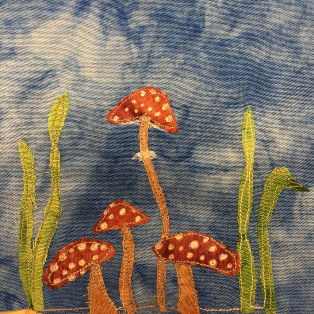 This quilt block shows four toadstools. These are the standard psychoactive toadstools featured everywhere, with orange stalks, red tops, and white spots. One toadstool is much taller and thinner than the others. There are also two stalks of grass and on either side of the toadstools.