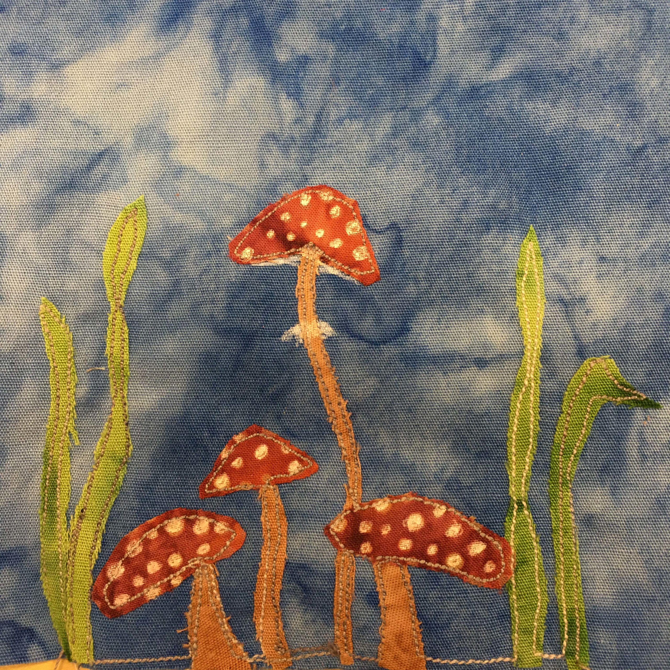 This quilt block shows four toadstools. These are the standard psychoactive toadstools featured everywhere, with orange stalks, red tops, and white spots. One toadstool is much taller and thinner than the others. There are also two stalks of grass and on either side of the toadstools.