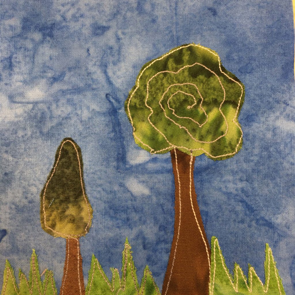 This quilt block shows jagged green grass at the bottom in a light green fabric. Then there are two trees juxtaposed, neither similar to the other. One tree is tall with a big round top of green (and spiral stitching) while the other is short and small with a triangle of dark grey green. These are both against a blue background.
