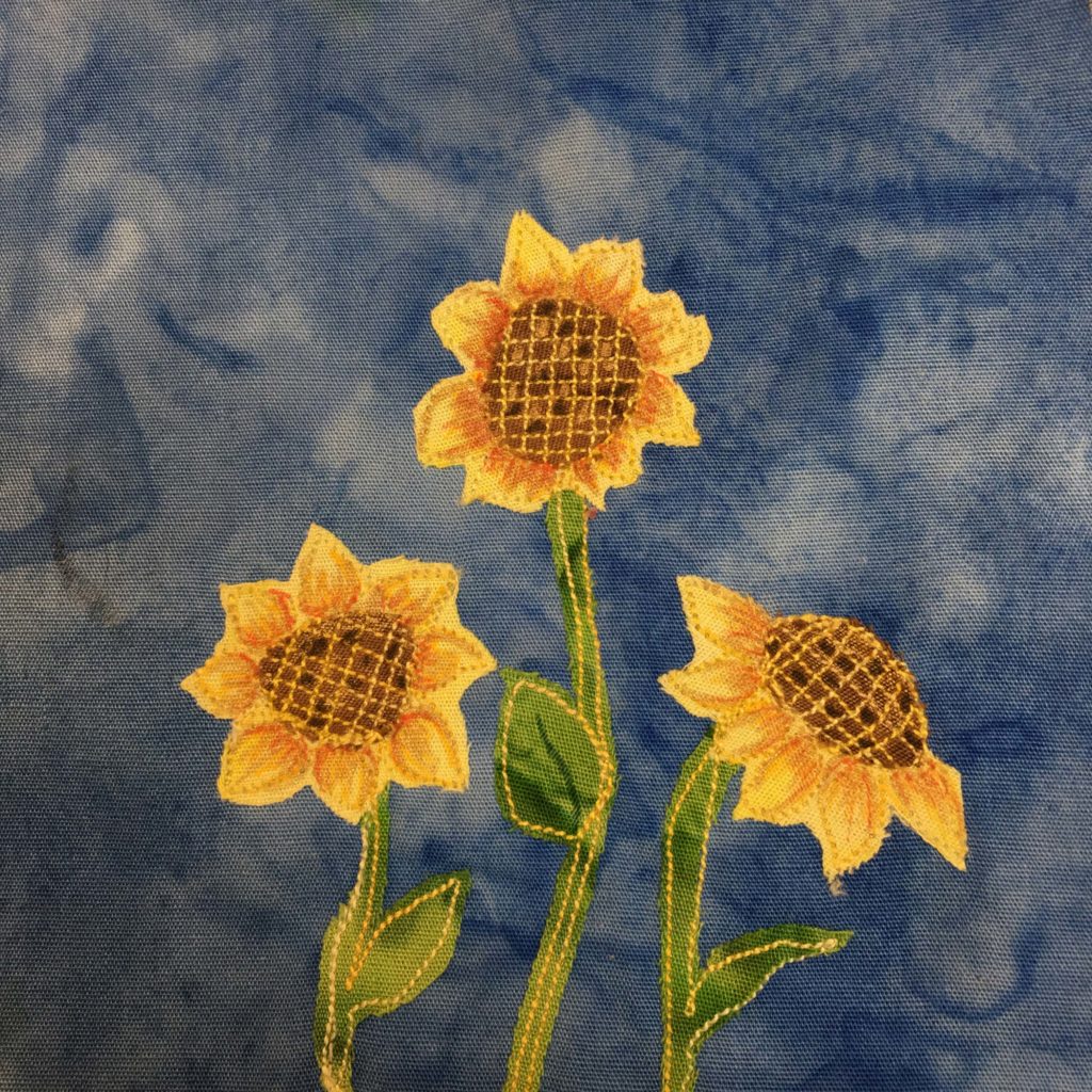 This quilt block has three sunflowers. Each sunflower has a hatched brown center with yellow shaded leaves surrounding it. It's quite detailed and beautiful. The leaves are shaded with an orange pen in a realistic fashion. The stalks are a green fabric with yellow stitched outlines and have a few leaves. This is against a slightly dark blue salmon-patterned background.