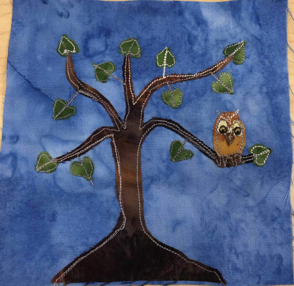 This quilt block shows a very dark brown tree with a big root base. There is no grass, but there are some dark green leaves on the branches of the tree, but not too many. On the right lower branch of the tree (there are 5) sits a brown owl. It has two large eyes, a triangular darker brown nose and little claws. The owl's details, including it's crown are made with a light colored stitching and a bit of yellow pen. All of this is against a moody blue salmon-patterned background.