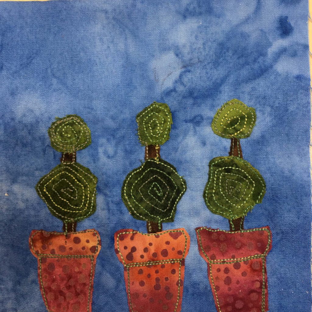 This quilt block shows three topiary plants in pots against a blue background. Each pot is a polka-dotted orange/maroon while the plants themselves are of one large ball and one small ball.  The greenery is a dark green with a spiral stitching inside.