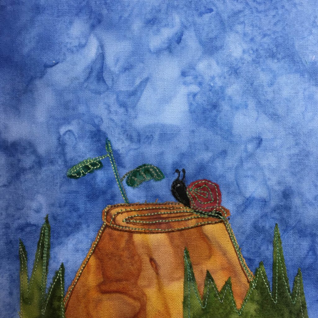 This quilt block shows a tree stump in some grass. There are dark green shades of fabric that are cut jagged to look like blades of grass at the bottom of the square. In the middle a orange-brown tree stump rises up. On top the tree stump, which is decorated with a swirling stitch is a black snail with a red shell. The snail is cresting the butte of the tree stump and looking to the left. It has two antenna. All of this is against a blue background.