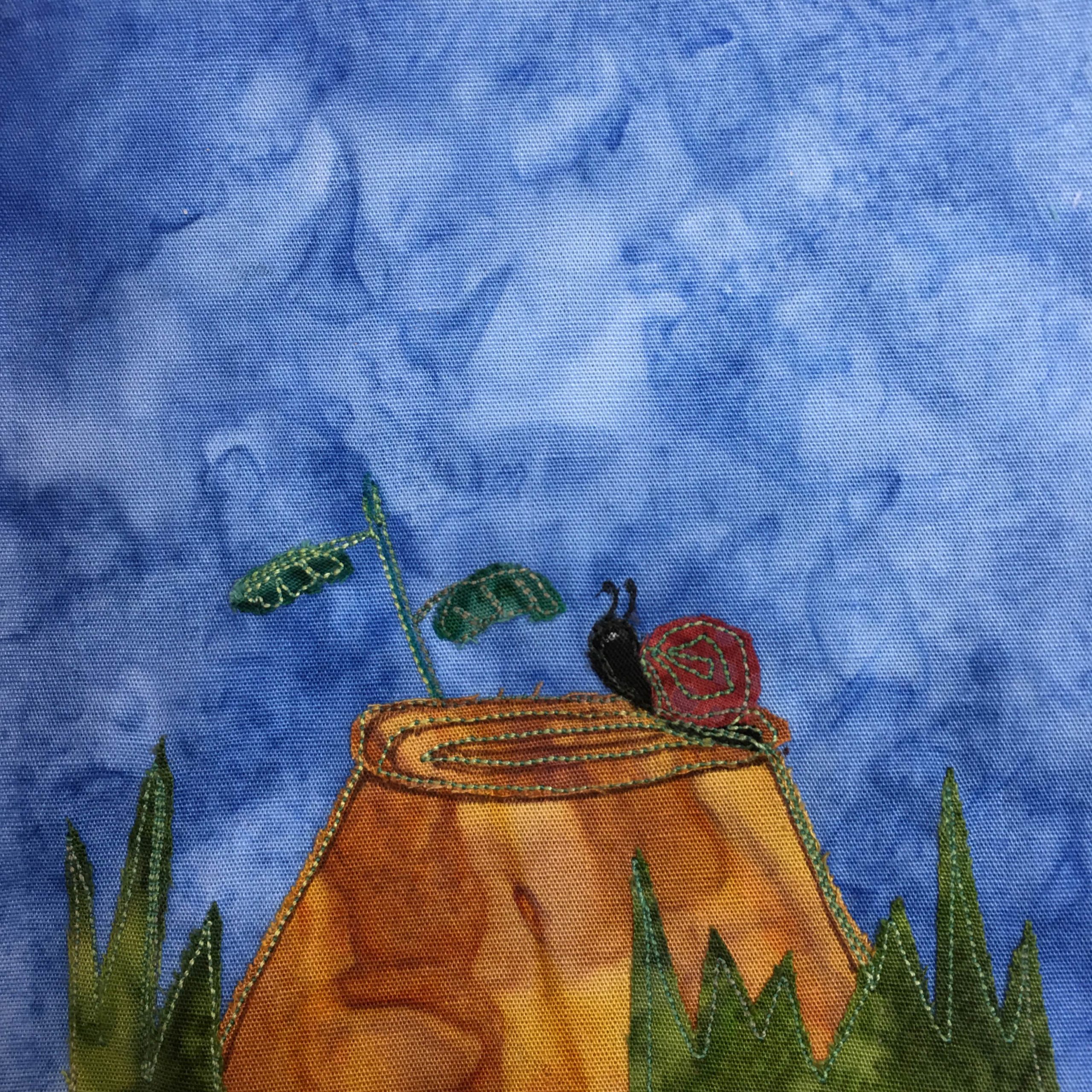 This quilt block shows a tree stump in some grass. There are dark green shades of fabric that are cut jagged to look like blades of grass at the bottom of the square. In the middle a orange-brown tree stump rises up. On top the tree stump, which is decorated with a swirling stitch is a black snail with a red shell. The snail is cresting the butte of the tree stump and looking to the left. It has two antenna. All of this is against a blue background.