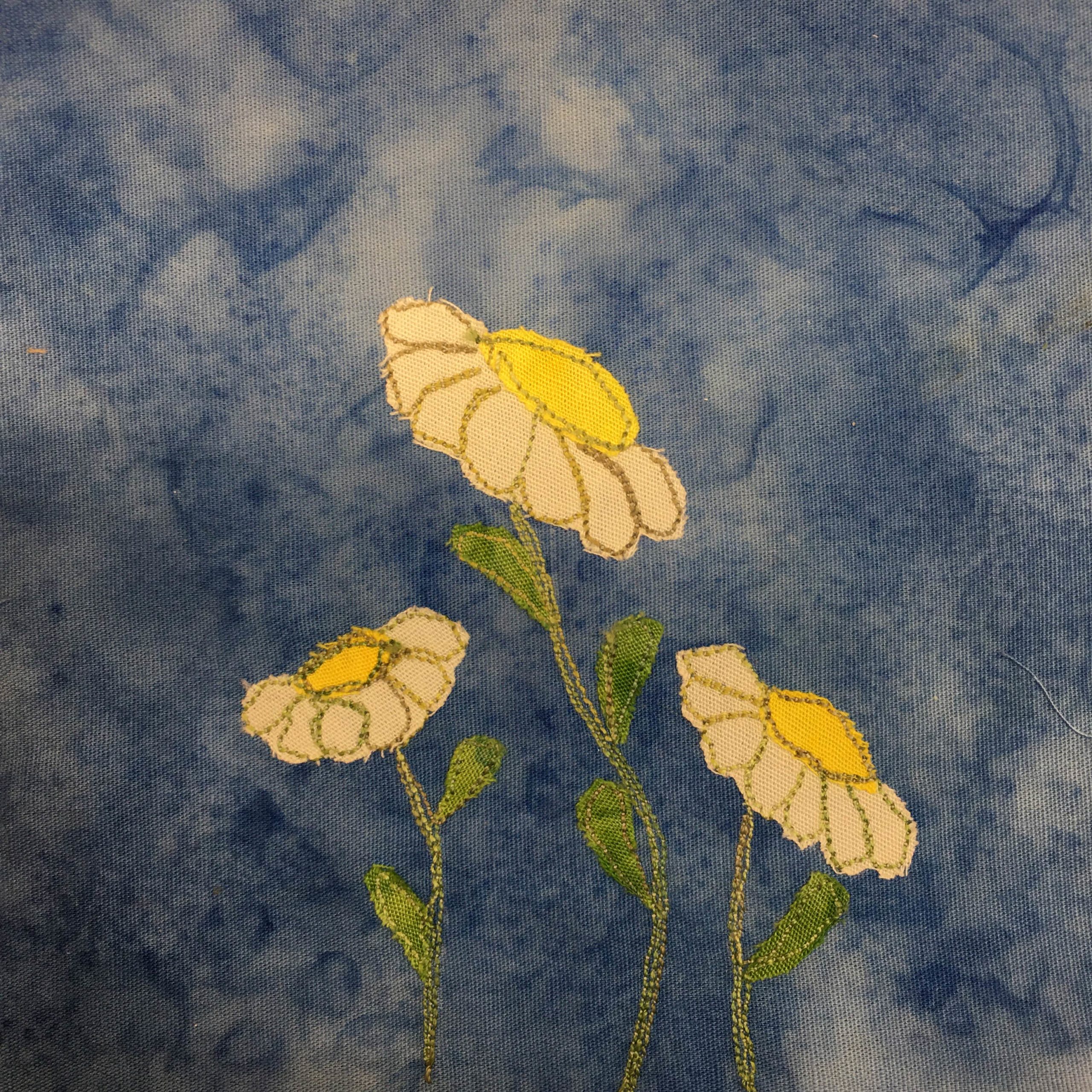 This quilt block shows Shasta daisies, three of them from left to right. The left daisy and right daisy are shorter and sit under the center taller daisy. Each one has a green stem with leaves. The top of each flower has a yellow center, and white petals.