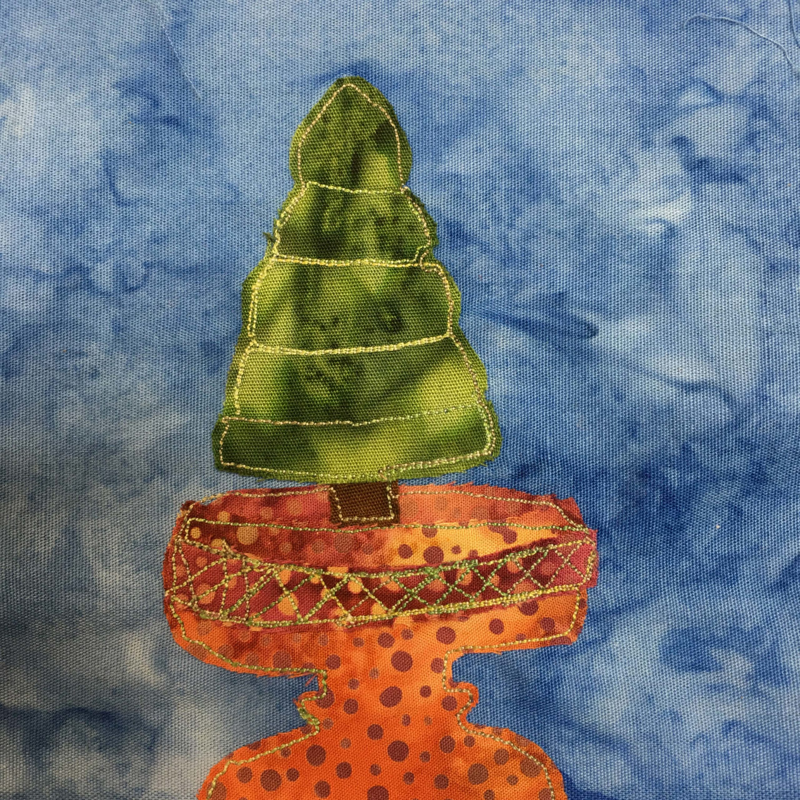 This quilt block shows an ornamental pot with a small spruce tree planted in it. The pot itself is a polka-dotted rust orange, with darker spots, and is shaped like a classic vase you would find in a greek garden. There is crossed stitches around its rim near the base of the tree. The small tree has stitches across it cutting it into sections as the green splotchy fabric tapers thinner towards the top. This is all against a blue salmon-patterned background.