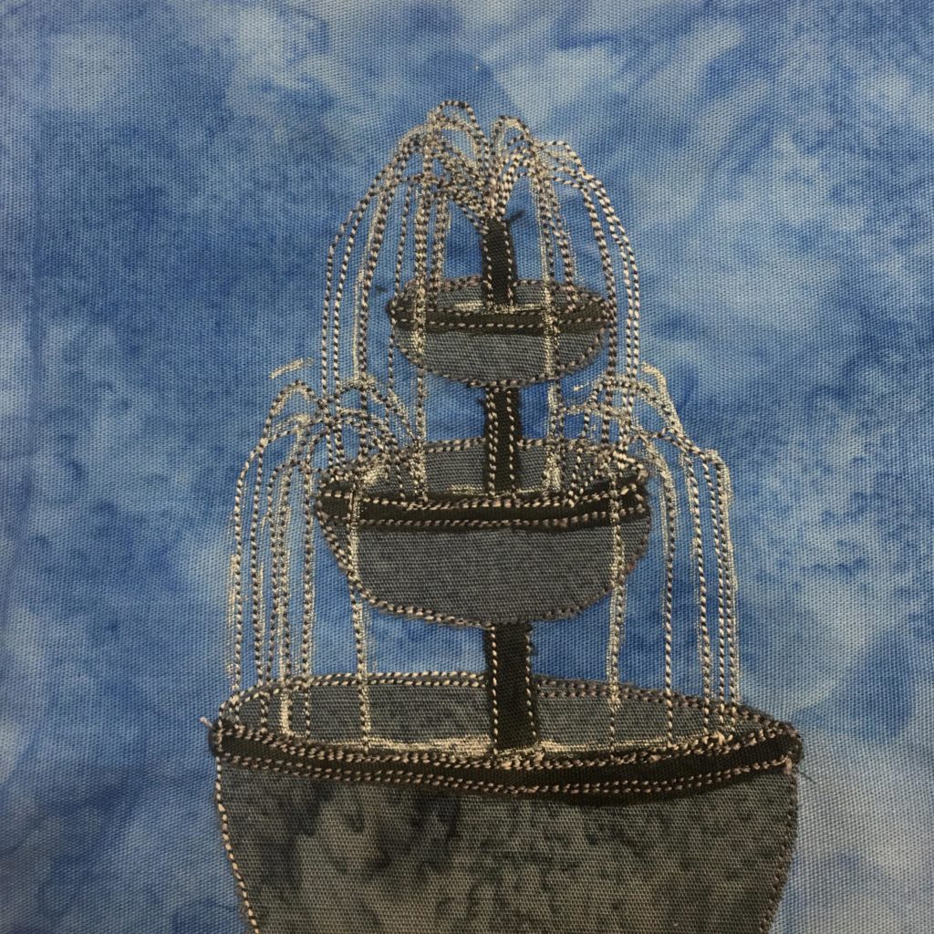 This quilt block shows a gray fountain against a blue salmon-patterned background. The fountain is in three levels, each level having a smaller gray bowl to collect the water. At the top of the central rod the water is shooting out and cascading down, all indicated by white and gray stitching lines. The gray bowls are lined at the top with a black fabric with additional stitching.