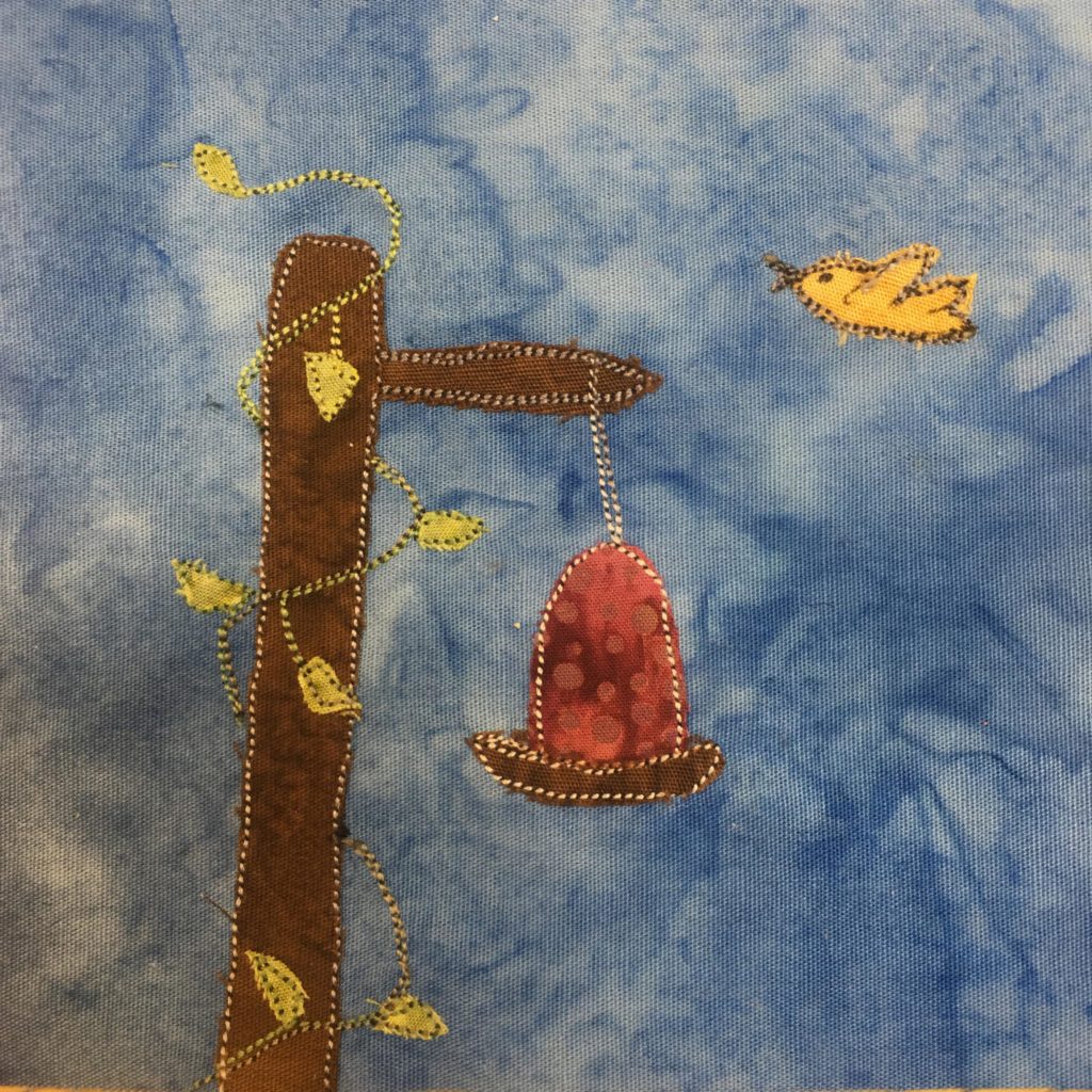 This quilt block shows a bird feeder hanging from a post against a blue background. The post is brown with a part jutting out that the feeder is hanging from. The post has a green vine made of green fabric and green stitching growing around and up the post. There is a yellow bird in the upper right flying. The feeder itself is red, like a hummingbird feeder, with polka dot pattern on it of another shade of red.
