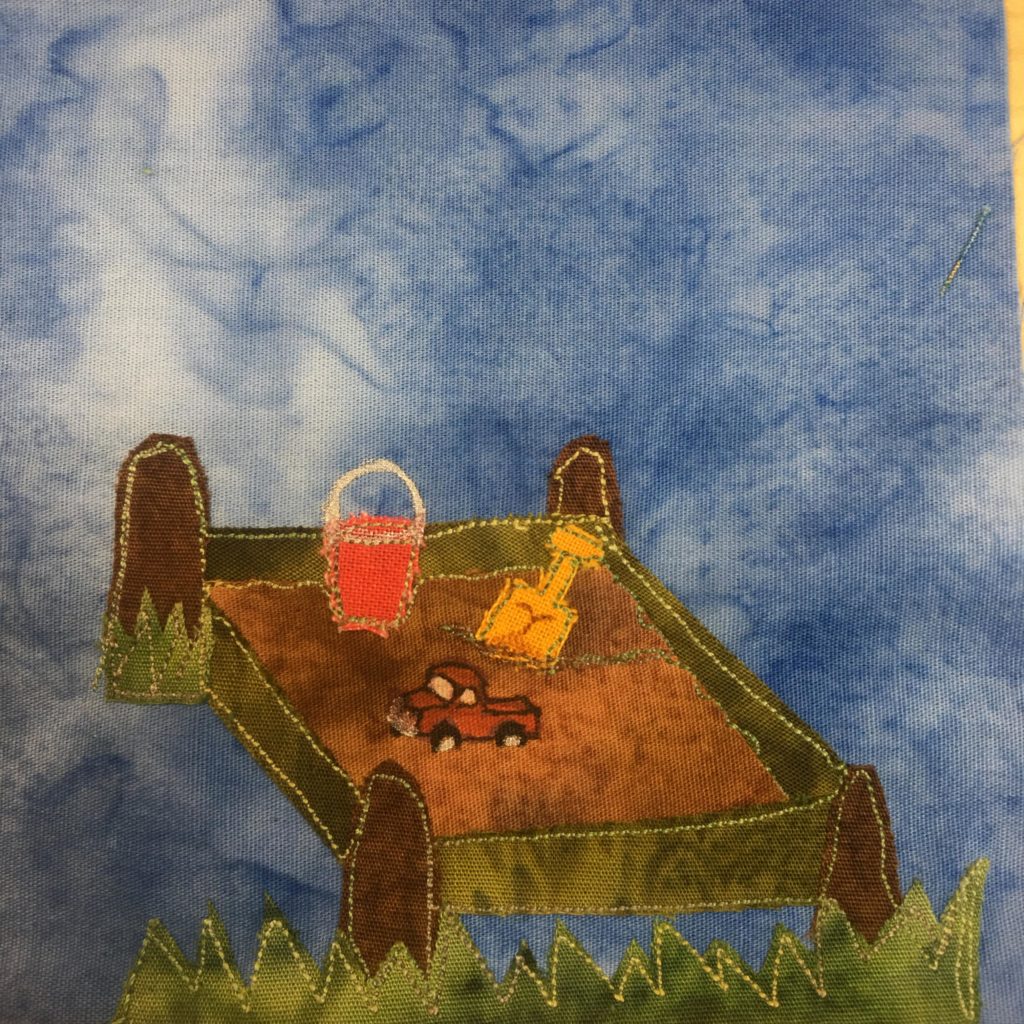 This quilt block shows a sandbox. Each corner of the sandbox is capped with the end of a brown log. The upper left brown log has grass at the bottom. The sides of the sandbox are made up of a green fabric indicating the walls/boundary. Inside is a light brown fabric for sand, a truck made out of pen, a yellow toy shovel stuck in the sand, and an orange bucket. At the bottom of the square is jagged grass. This is all against a blue salmon-patterned background.
