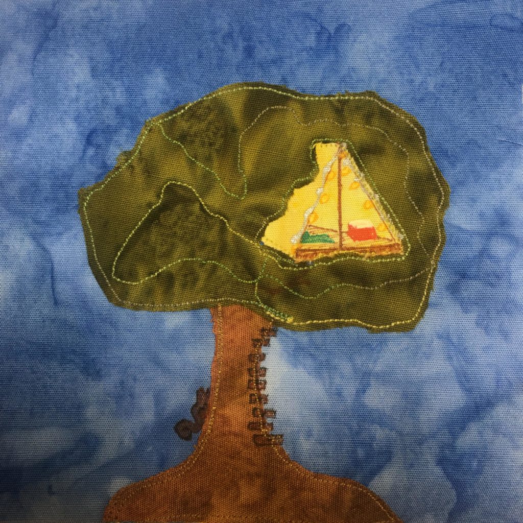 This quilt block shows a very large tree with a tree house inside the greenery. On the side of the brown trunk is a squirrel and a bunch of steps for climbing the tree.  The greenery is an amorphous green fabric with green stitching around it. In that amorphous shape is a yellow tent-like shape with various items inside and lights along the edges. The tent is the tree house. This is all against a darker blue background.
