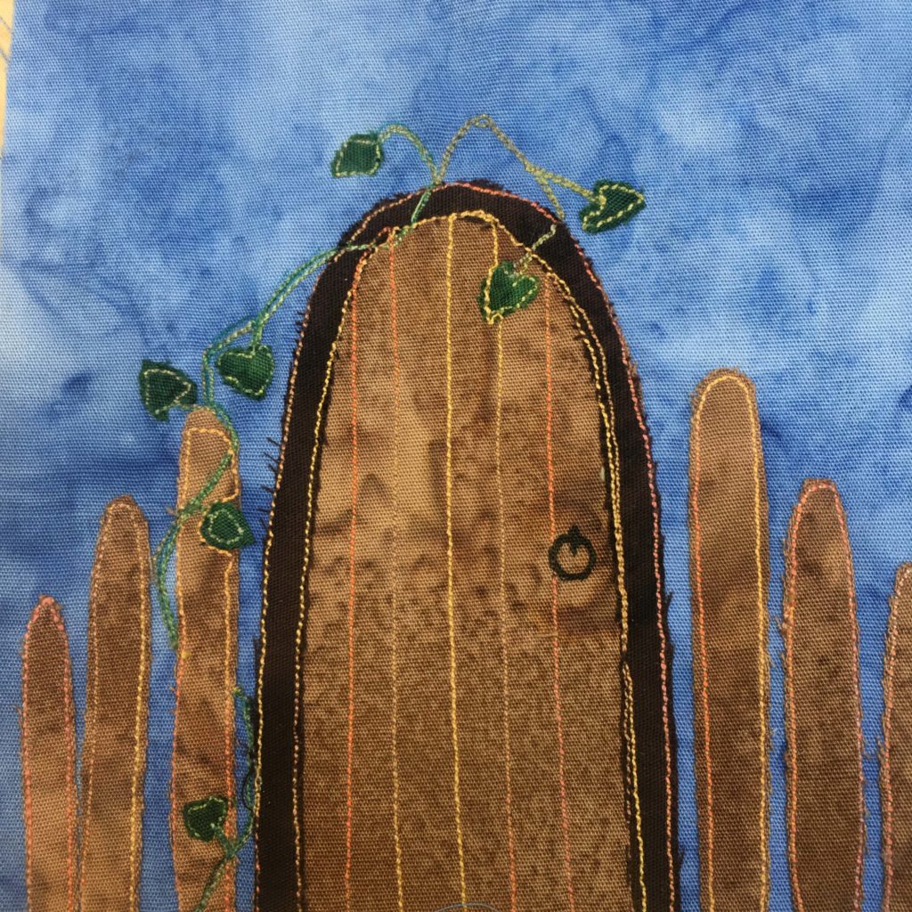 This quilt block shows a large wooden door with a big metal ring as a door handle. The door is actually a gate, and is framed by a much darker brown outline of fabric. Next to the door are fence posts that decrease in height as they move towards the edge of the block. There is a green vine made of dark green fabric and green stitching that travels up the post and dangles over the top of the door. The gate or door itself is a light brown with multicolored threading indicating the slats of the wood. This is against a blue salmon-patterned background fabric.