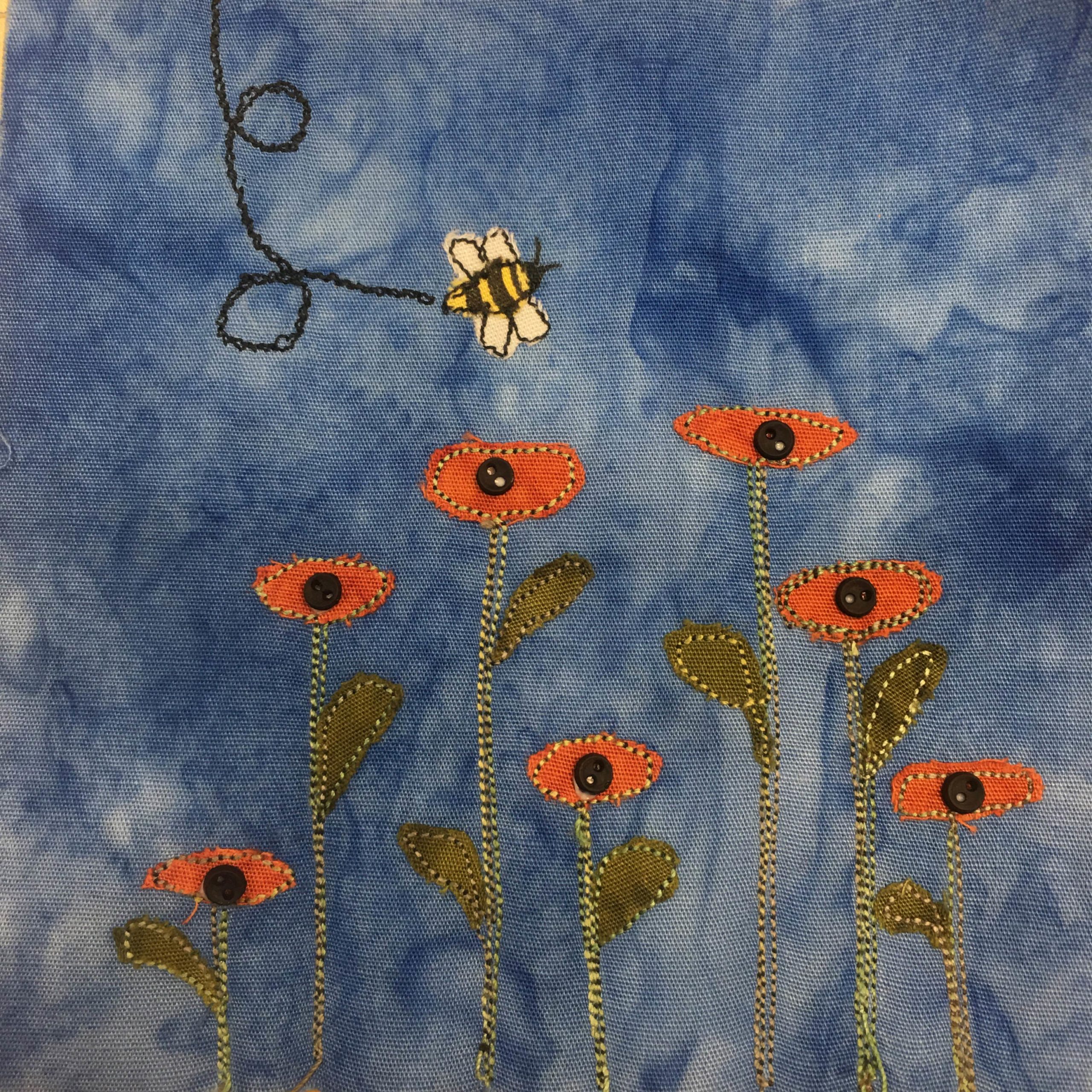 This quilt block shows flowers in a row of varying heights, and above them a wandering bee. The bee's trail is a black stitching, and the bee itself is made of yellow and white fabric. Stripes are penned in for the bee's appearance. The flowers are straight stalks of stitching with some dark green leaves poking out the side. At the top of each stalk is an eye shaped piece of orange fabric, and in the center of those eyes is a small black two holed button. This really makes them look like eyes!