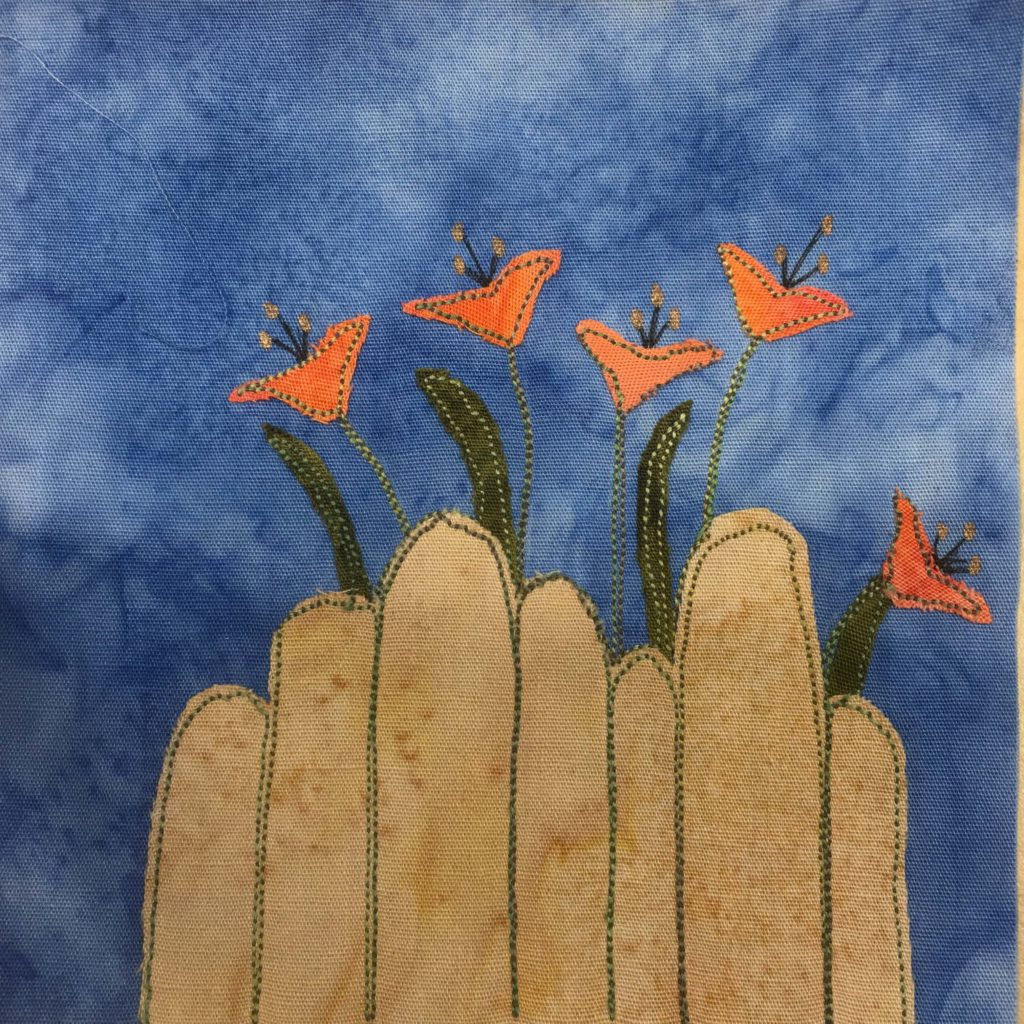 This quilt block shows a light brown slatted fence with curved (round) ends with day lily flowers behind it. The flowers are made out of light orange fabric for the bells/blooms and have little pen lines coming out of them for the pistels. There are also long green leaves too. The flowers and leaves (and fence) are outlined with a green stitching. This is all against a blue salmon-patterned background.