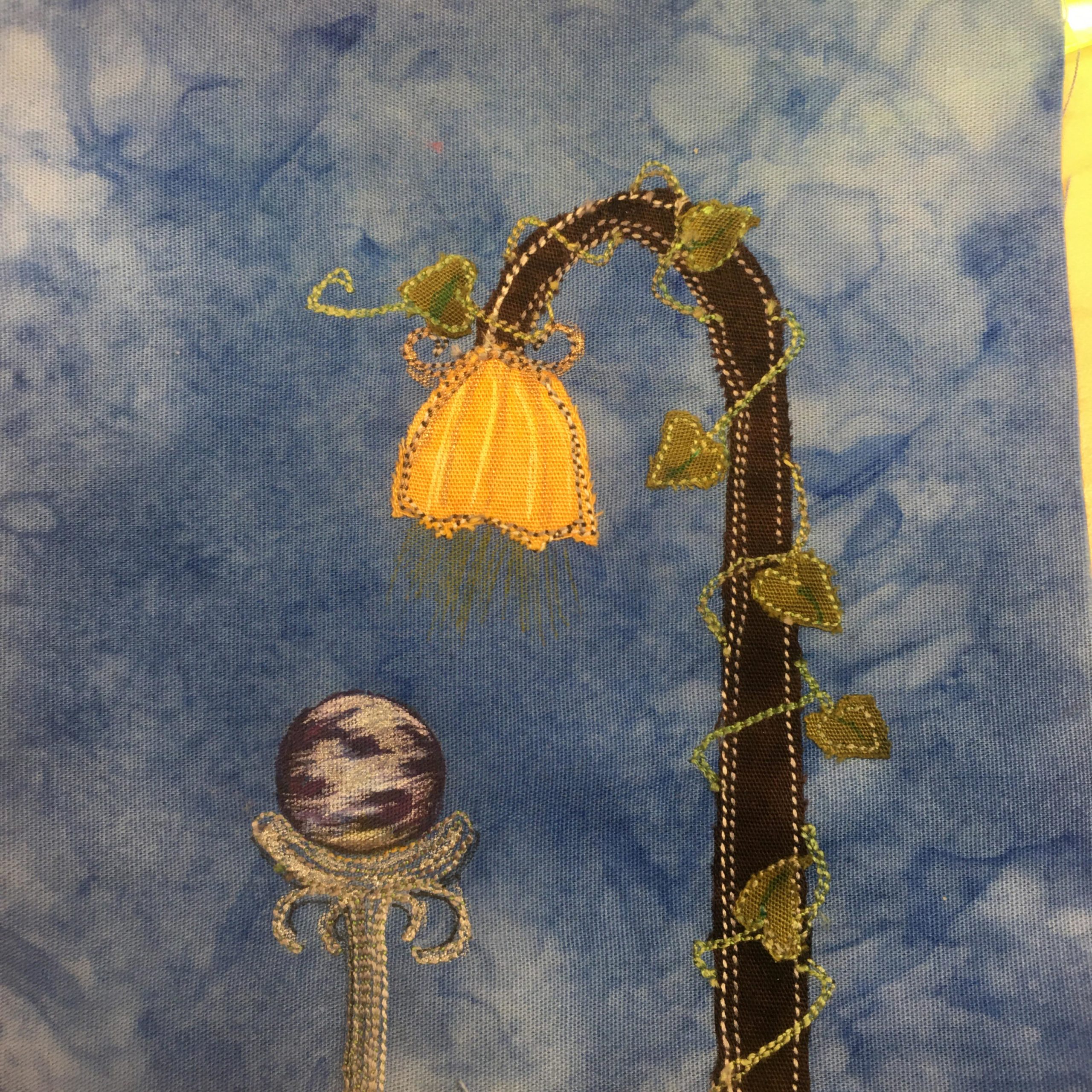 This quilt block shows a shephard's crook with a vine around it attached to a yellow bell flower shaped lampshade. Inside the lamp shade is a light that's shining out with yellow pen marks showing the light. Below the bell flower light is a pedestal sort of stick with a purple mystical ball on top. It's a garden gazing ball and it's been marked with pen to make it look very mysterious. The pedestal is made up mostly of white stitching, while the crook and lampshade are a dark brown and bright yellow. The vine is green with little leaves.