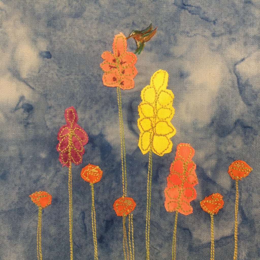 This quilt block shows a variety of colors of hollyhocks. The stalks of the flowers are green stitches, while the bodies of the flowers are different colored fabrics: orange, maroon, and yellow. One flowe rbody has pen marks for the pistles, and on that flower there is drawn a beautiful hummingbird. The hummingbird is drawn with different color pens with its wings out.