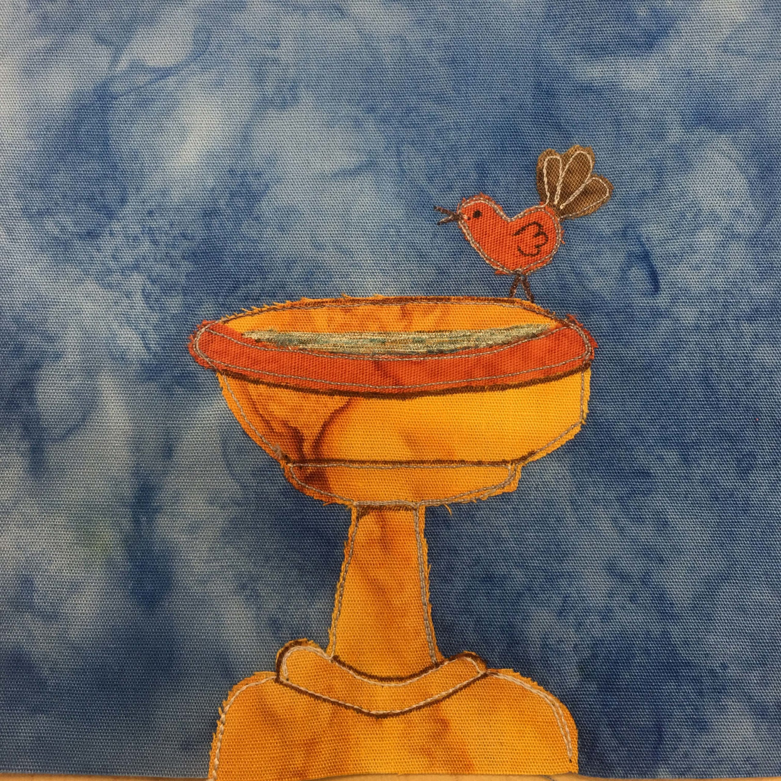 This quilt block shows a golden bird bath with a little bird on it. This is against a blue salmon-patterned background. The birdbath has a wide circular base, a cylinder support, and then a large concave half globe holding a bit of water. The bird is a deeper orange with dark tan tail feathres. The birdbath is outlined by dark stitches and light stitches, and has a bit of pen indicating water.