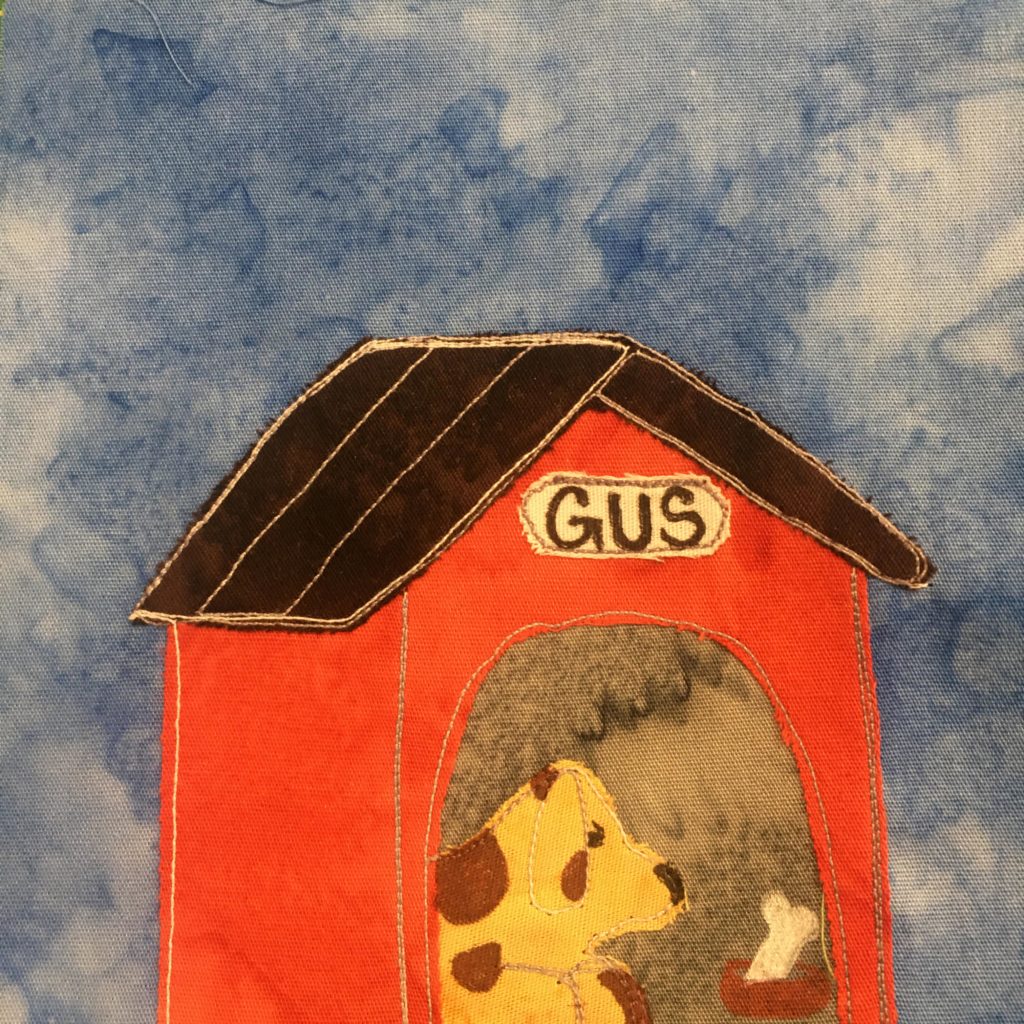 This quilt block shows a dog house against a blue salmon-patterned background. The dog house has a very dark brown roof and is made of bright orange fabric. The inside is a gray fabric, but also inside is a spotted yellow dog. The dog is yellow with brown spots on it. There is also a dog bowl with a white bone in it. The bowl and bone are drawn with a pen.