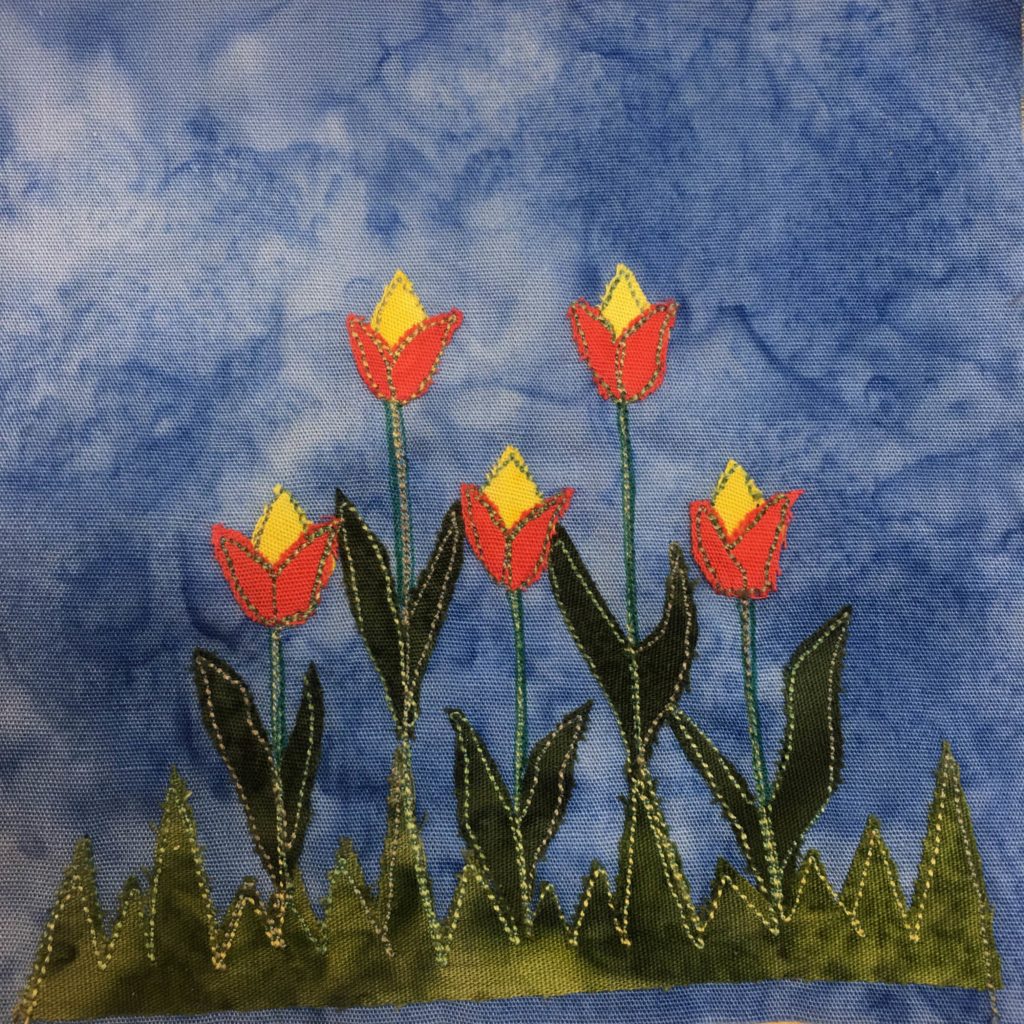 This quilt block shows a bunch of flowers. Five tulip looking flowers extend upwards from a jagged piece of green fabric made to look like grass. The stalks of the flowers are pen and stitching. The leaves that point upwards, two for each flower on each side are a darker green with a light green stitching outline. The tops of the flowers, the bulbs, are a bright orange at the base, with a yellow part sticking out the top, much like a tulip.