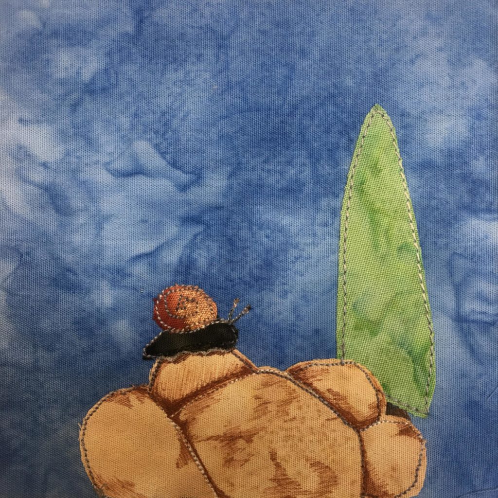 This quilt block shows a small pile of rocks with a small (but tall) bush next to it in the shape of a tree. On the pile of shaded (with a pen) tan rocks is a black snail. the snail shell is an ombre rust and tan with a spiral stitching inside it. The mollusk is black with two antenna. The bush/greenery is almost as tall as the block and kinda pointy. This is all against a blue salmon-patterned background.